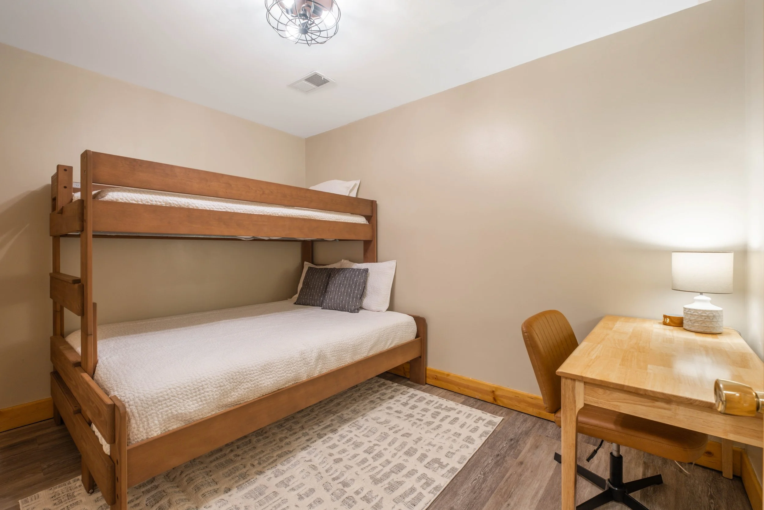 Small bedroom with wooden bunk beds, a desk with a lamp, and a beige carpet.