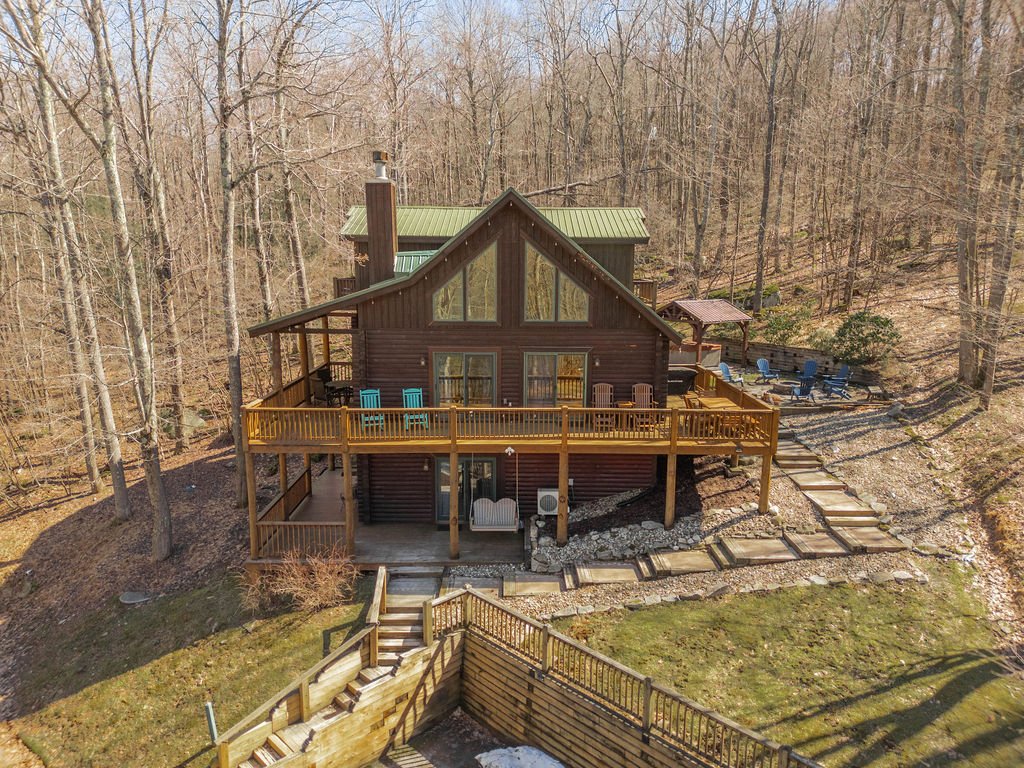 A brown multi-story house with large angled windows and a green metal roof, surrounded by trees, with a spacious wooden deck and outdoor seating area on a hillside.