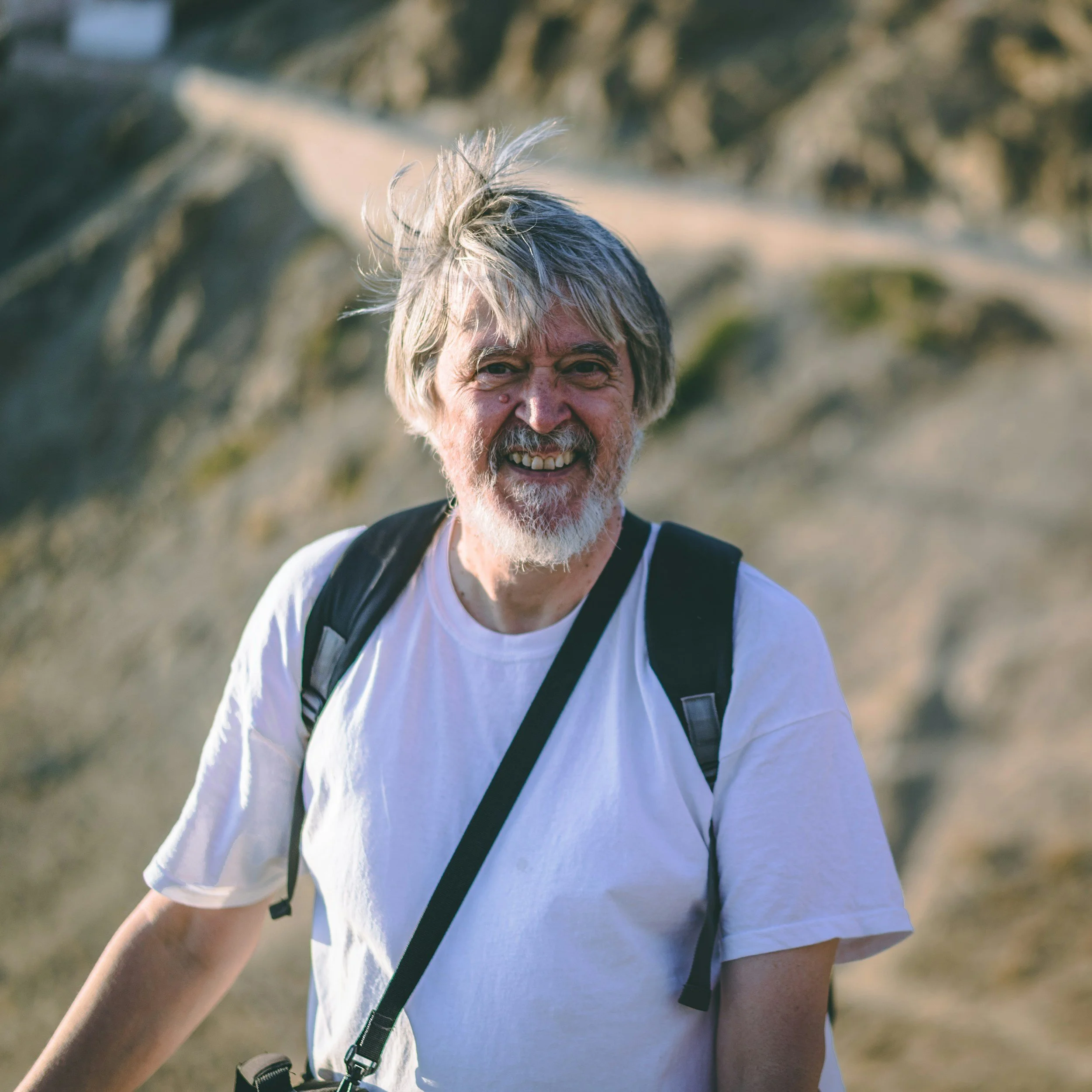 Close-up of a smiling man with gray hair and beard, wearing a white t-shirt and a black strap, outdoors with blurred natural background.