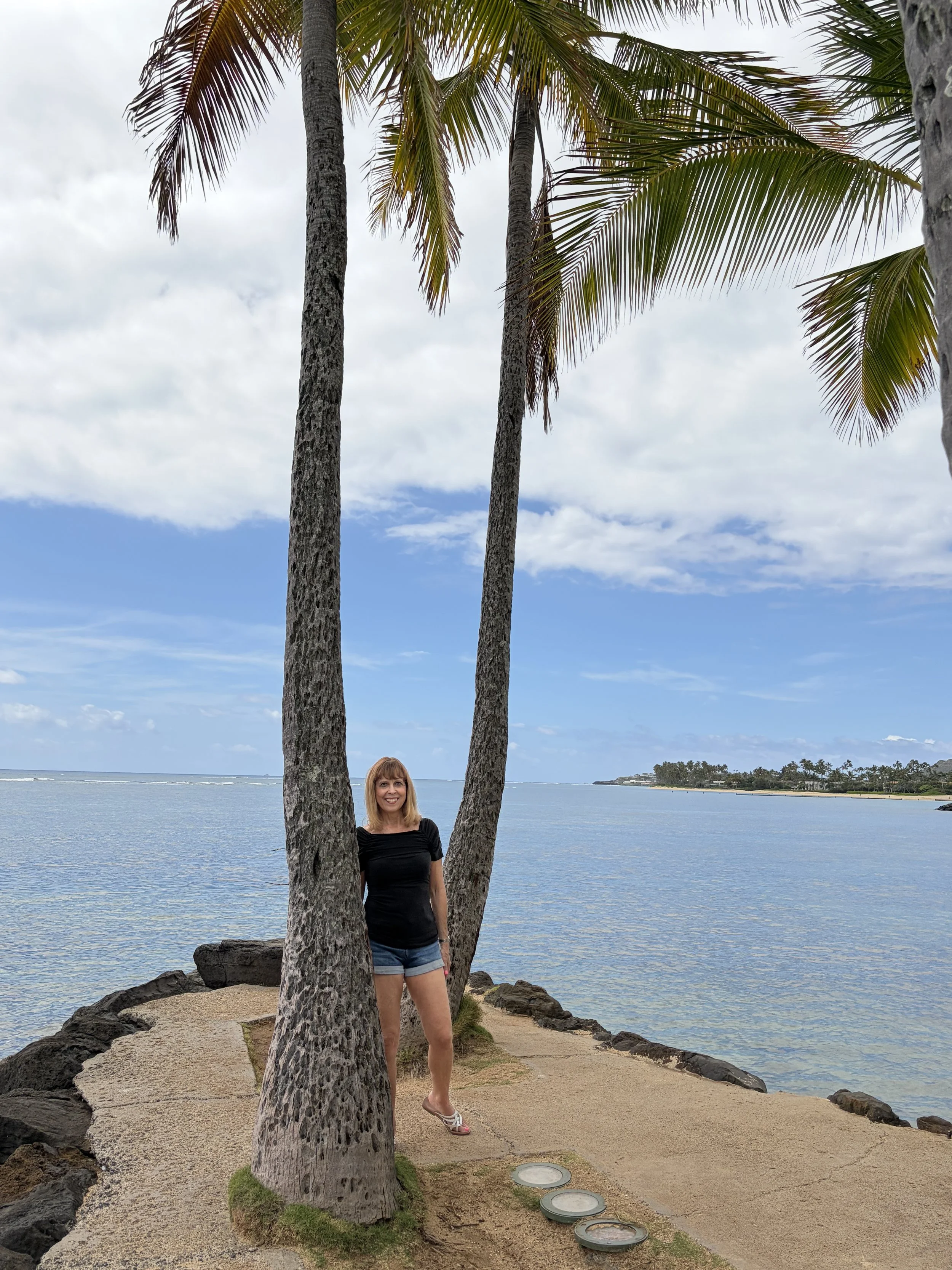 A woman standing on a beach near two palm trees, with water and a distant shoreline in the background.