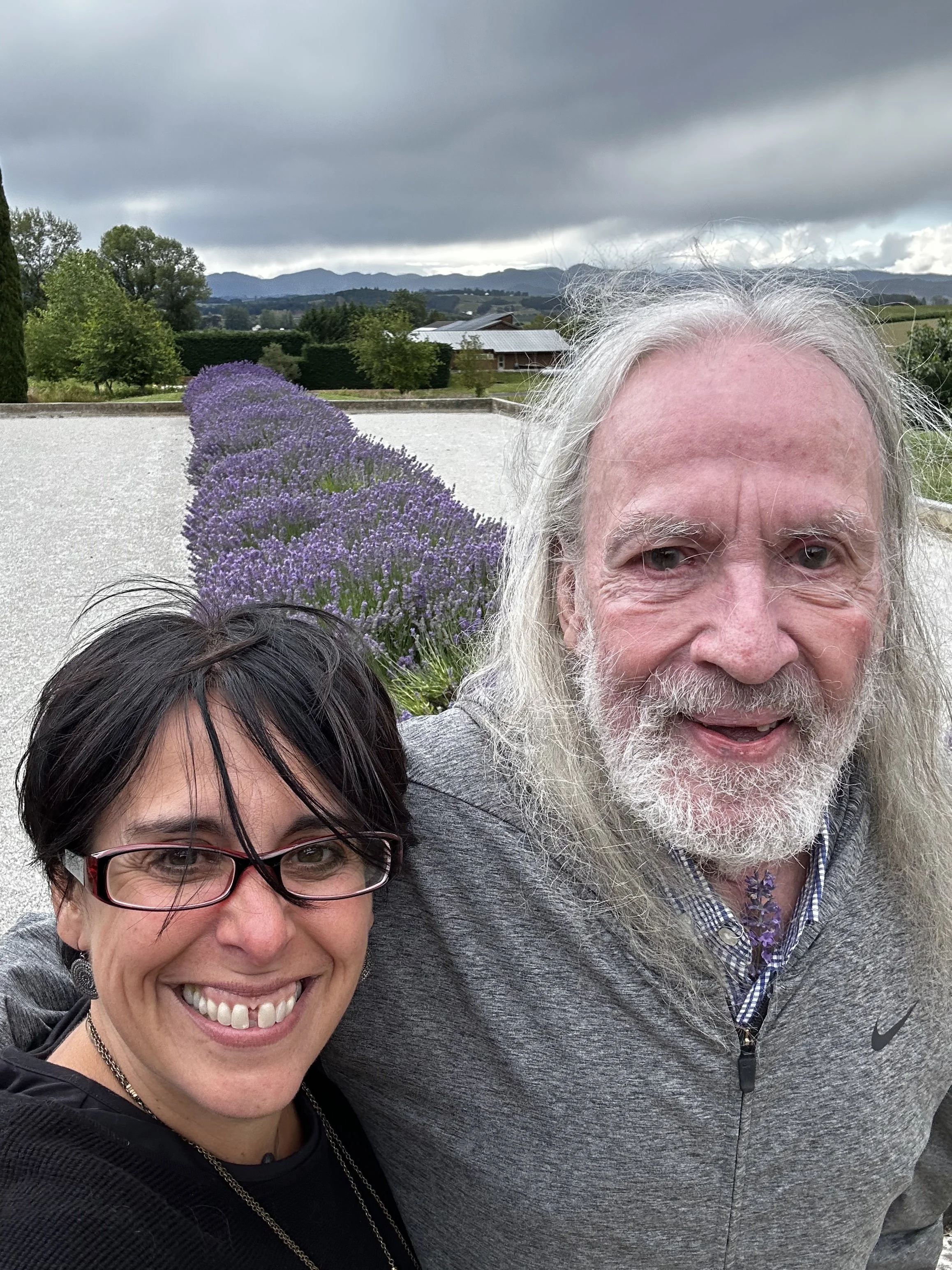 Meka and her father smiling, taking a selfie outdoors with a background of purple lavender flowers, trees, a building, mountains, and a cloudy sky.