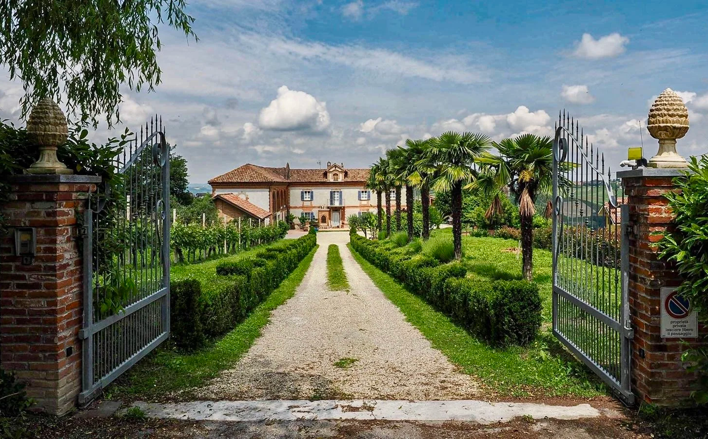 Beautiful winery in Piedmont, Italy. A gravel driveway leading to a large house, flanked by trimmed bushes and palm trees, with a wrought iron gate and brick pillars at the entrance, under a partly cloudy sky.
