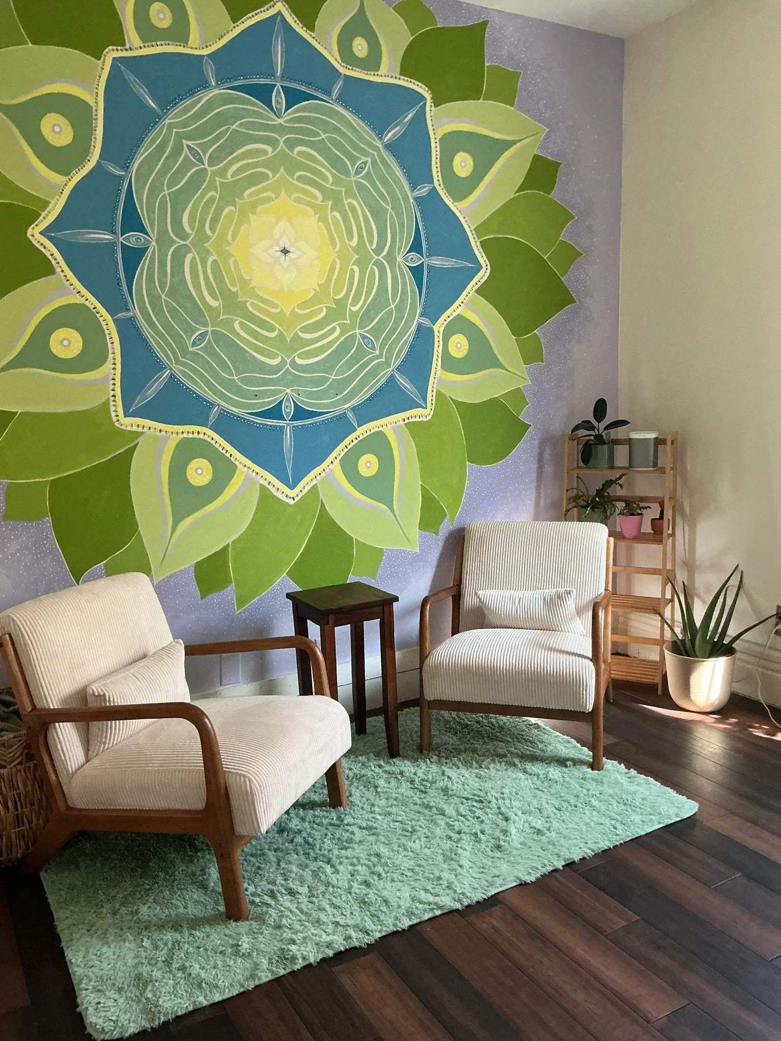 A cozy living room corner with a cactus green shag rug, two cream armchairs with wooden frames, a small black side table, and a wooden plant stand with potted plants. The wall features a large, colorful mandala-style painting with green, blue, and yellow hues, and a purple background.