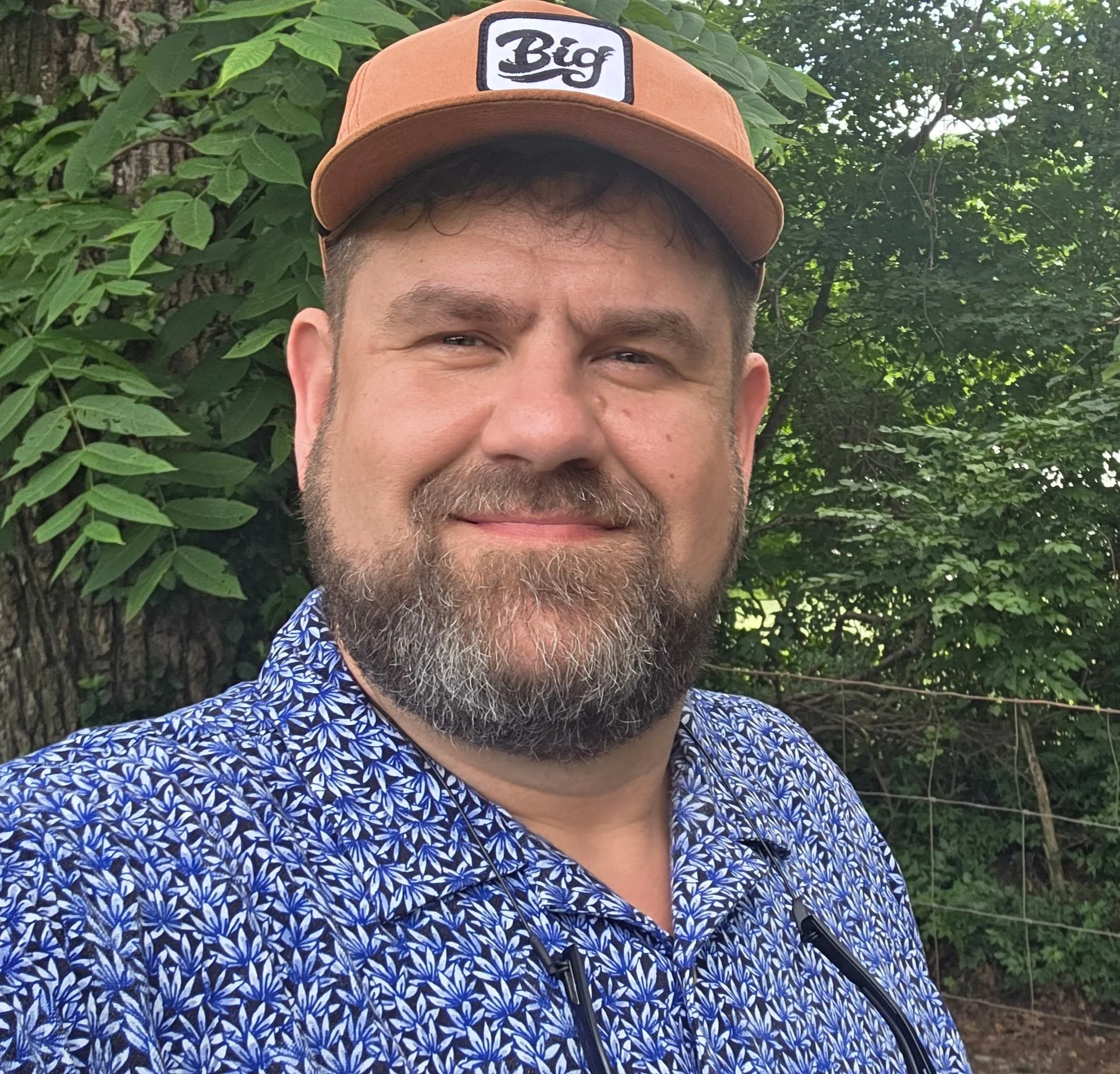 A man with a beard and mustache smiling, wearing a brown cap with a 'Big' patch on it and a blue patterned shirt, standing outdoors in front of green leafy trees.