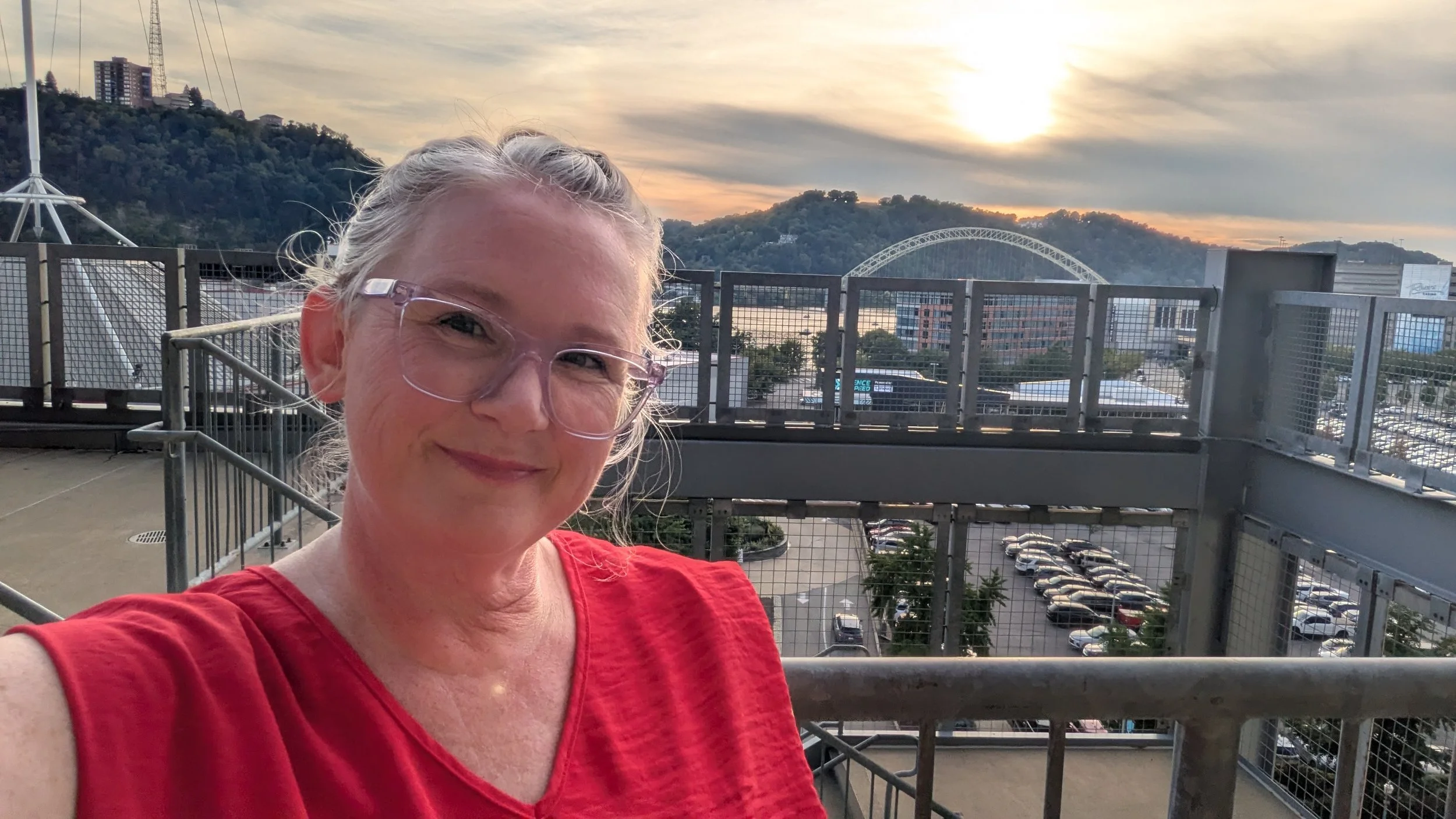A smiling woman with gray hair and glasses taking a selfie on a rooftop parking deck during sunset, with a cityscape and hills in the background.