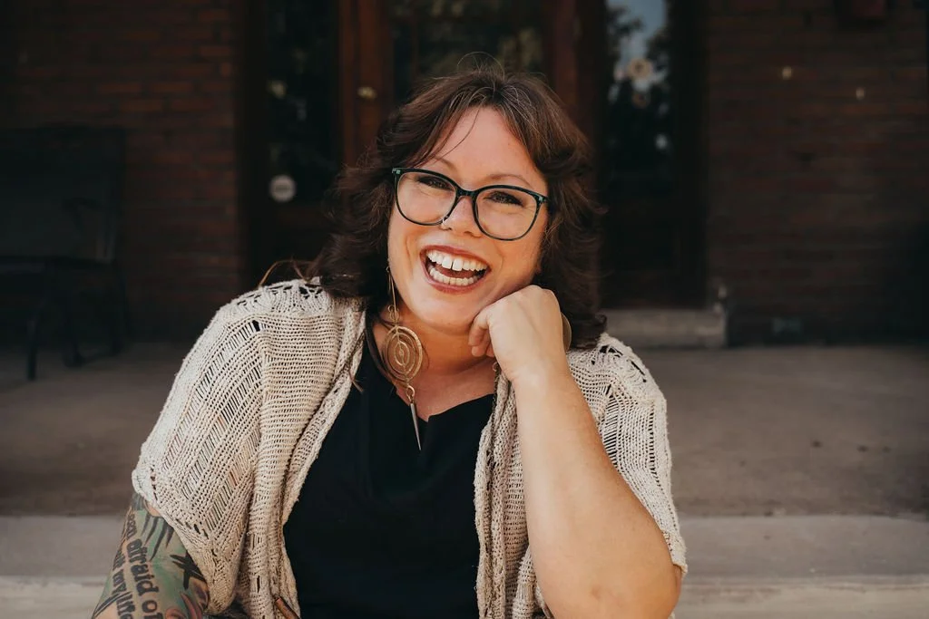 A woman with glasses and dark wavy hair smiling outdoors, resting her chin on her hand, in front of a brick building.