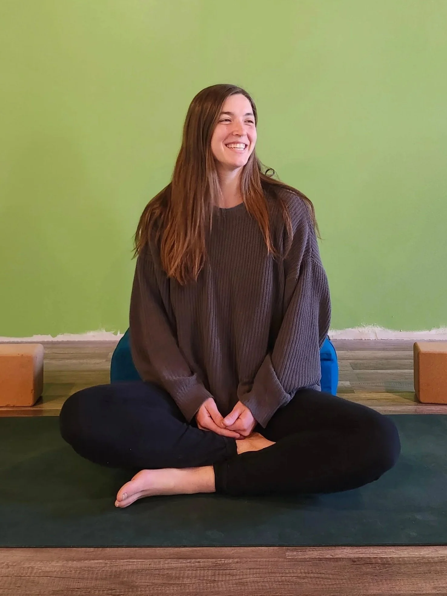 A woman sitting cross-legged on a yoga mat, smiling, with a green wall behind her and a yoga block beside her.