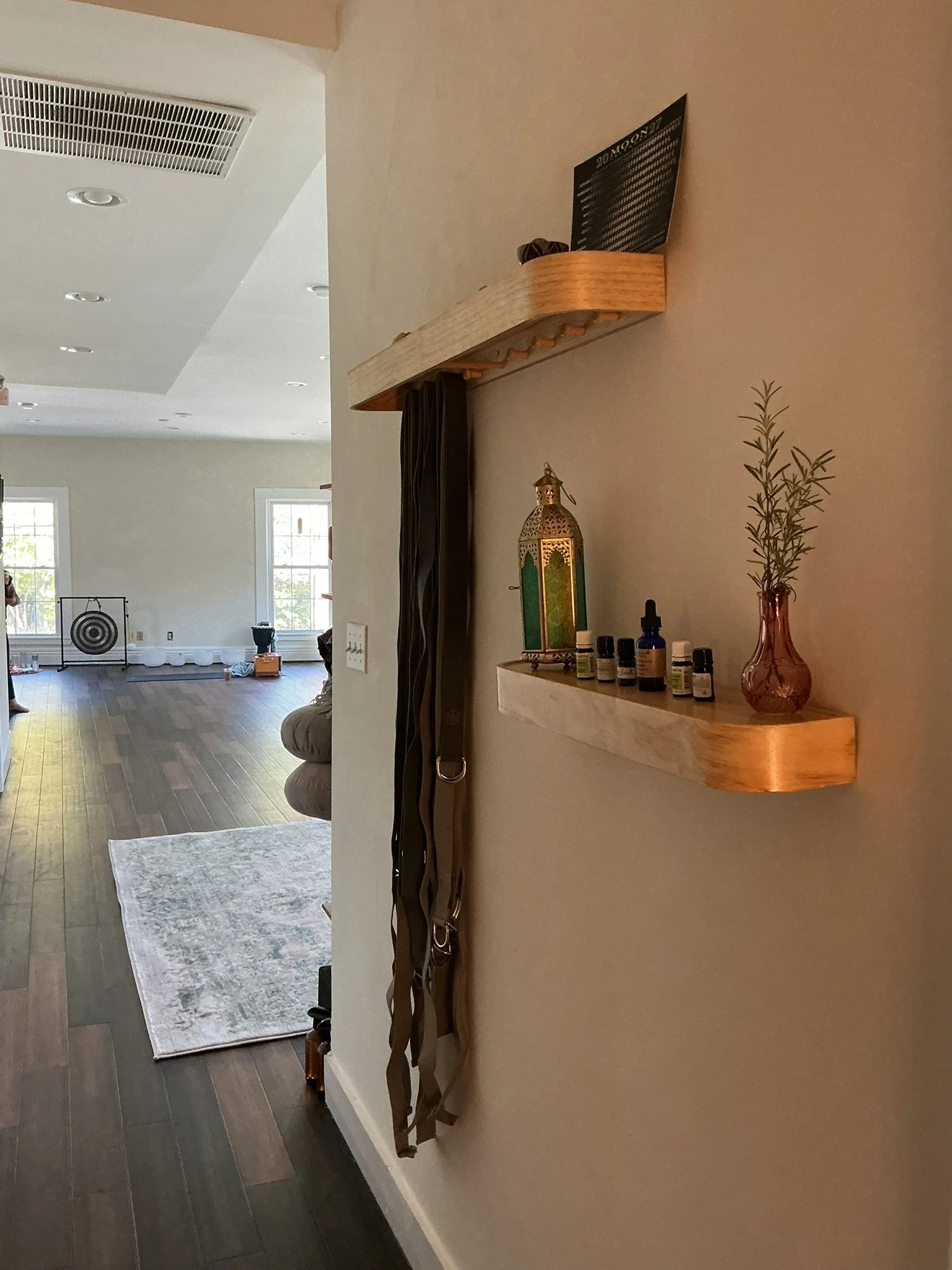 Interior view of a living space with a wooden wall-mounted shelf holding a decorative lantern, drops of essential oils, and a vase with tall plant stems. In the background, a rug, windows, and part of a person are visible.