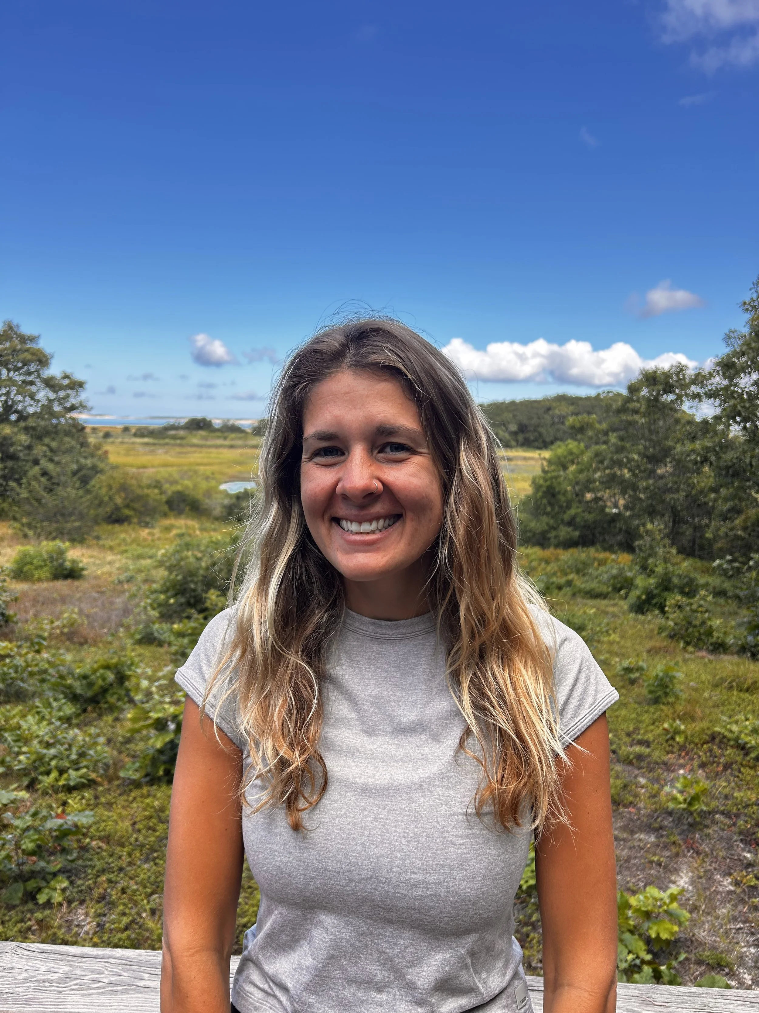 Close-up of a smiling woman with long wavy hair standing outdoors in a natural landscape with trees, bushes, and a bright blue sky with scattered clouds.