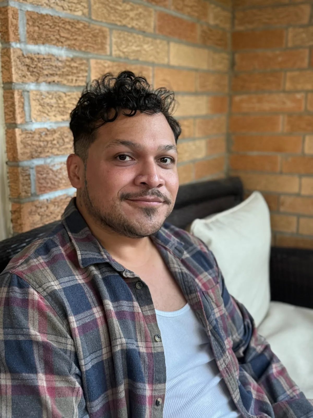 A man with curly dark hair, a light beard, and a slight smile wearing a plaid shirt over a white tank top sitting on a sofa with a brick wall background.
