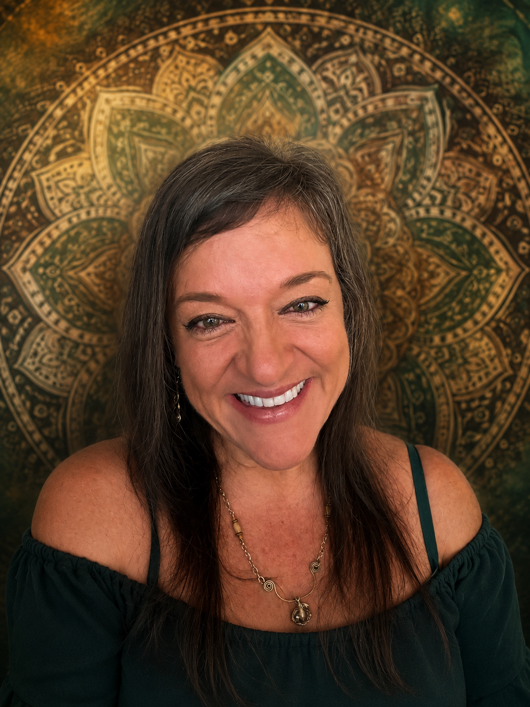 A smiling woman with shoulder-length brown hair, wearing a dark off-shoulder top and a gold necklace with a pendant, standing in front of a detailed gold and green mandala wall hanging.