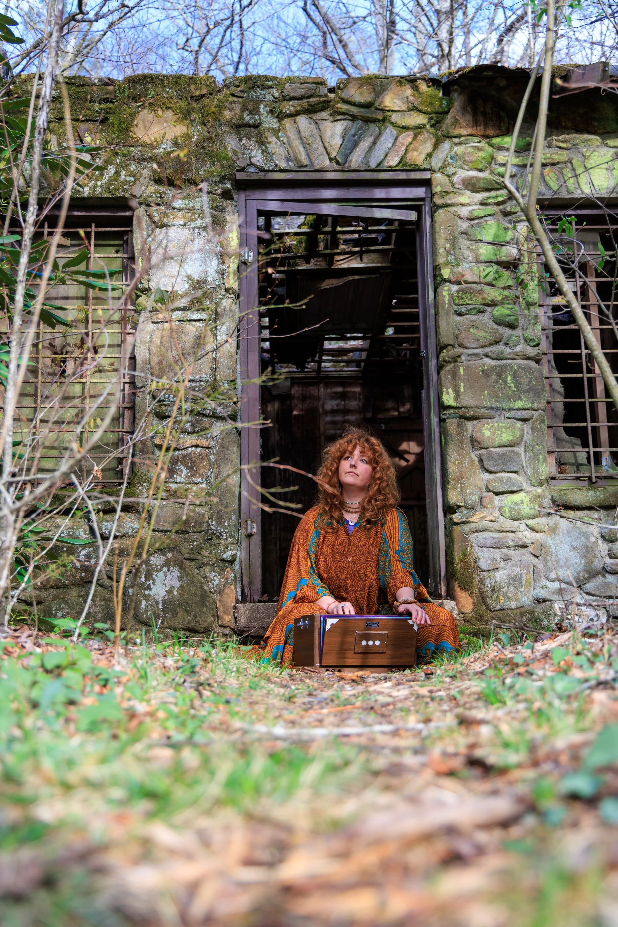A woman with curly red hair, wearing a colorful, patterned dress, sitting outside in front of an old, abandoned stone building. She is holding a wooden box or instrument in her lap, and inside the building, the roof is partially collapsed.