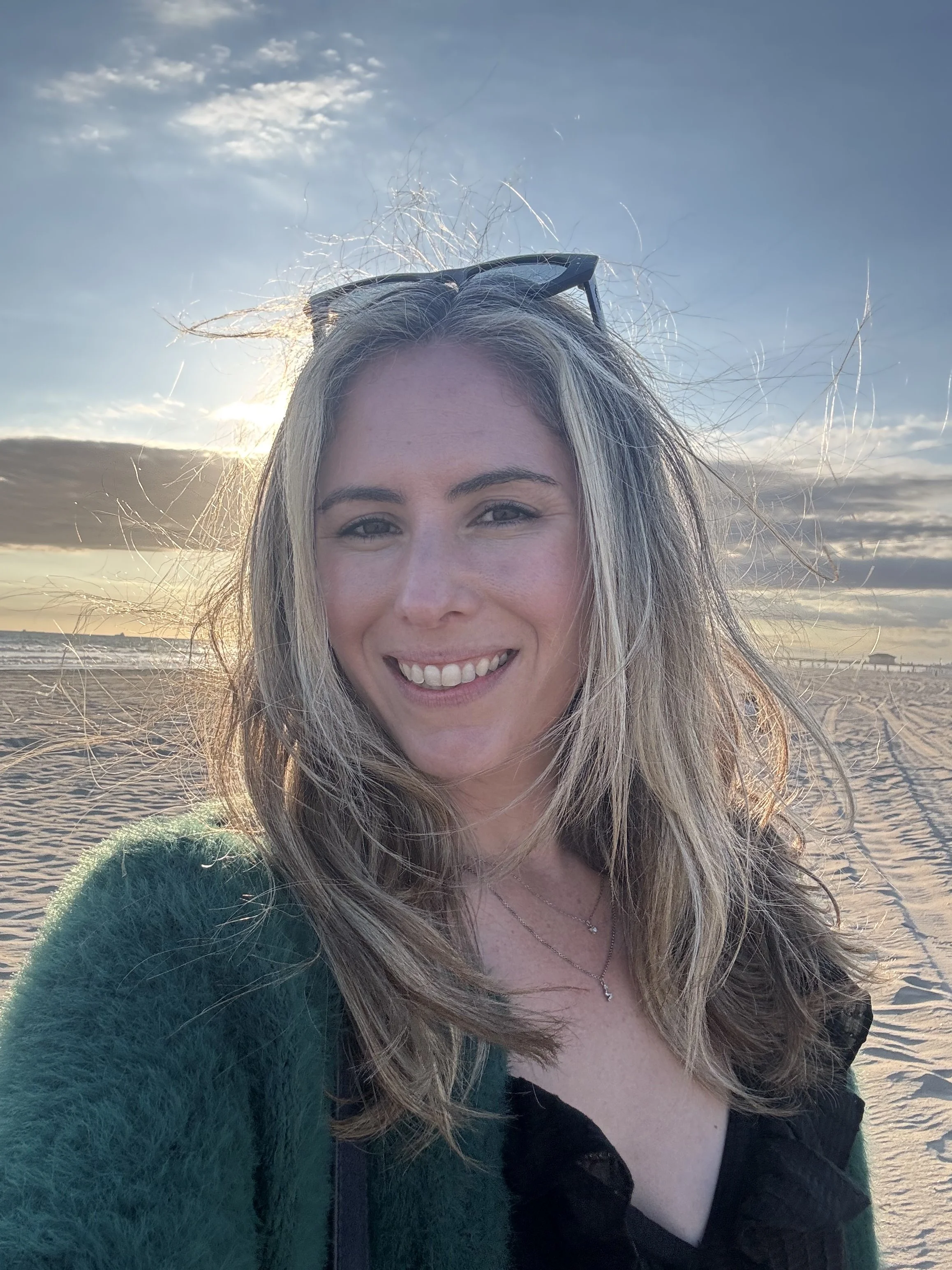 Close-up of a woman with long blonde hair smiling at the beach during sunset, wearing a green fuzzy sweater and a black top, with sunglasses resting on her head.