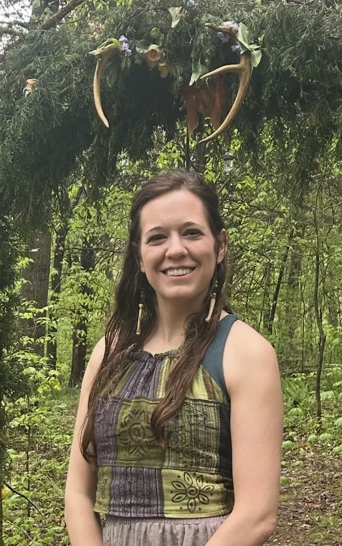 A smiling woman with long hair wearing a patterned sleeveless top standing in a forested area with green foliage.