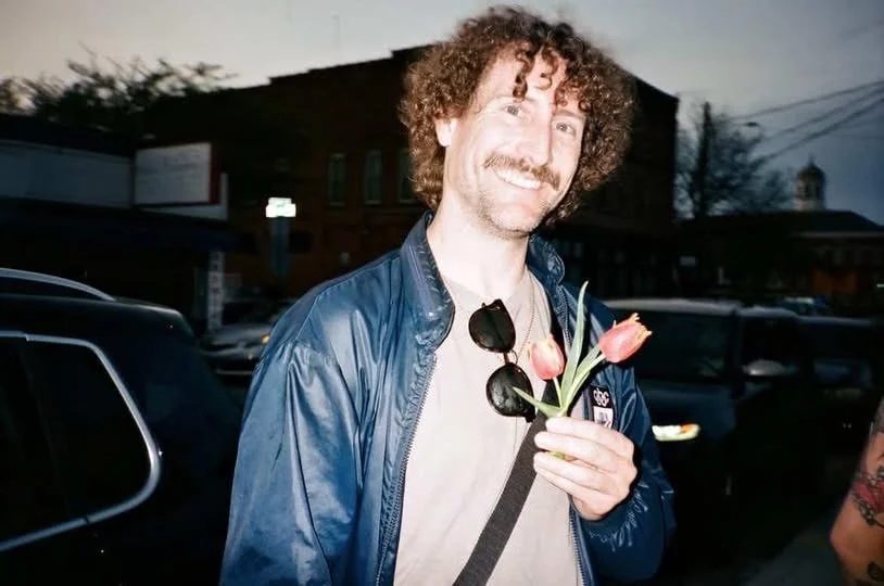 A man with curly hair and a mustache smiling outdoors, holding pink tulips, wearing a blue jacket and sunglasses hanging from his collar, in a parking lot at dusk.