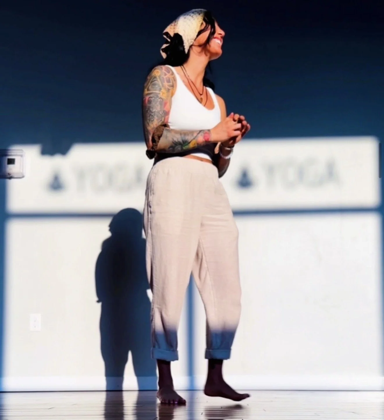 A woman standing barefoot on a wooden floor, smiling, with dark hair, tattoos on her right arm, wearing a white crop top, beige pants, and a patterned headscarf, silhouette cast against a bright background.