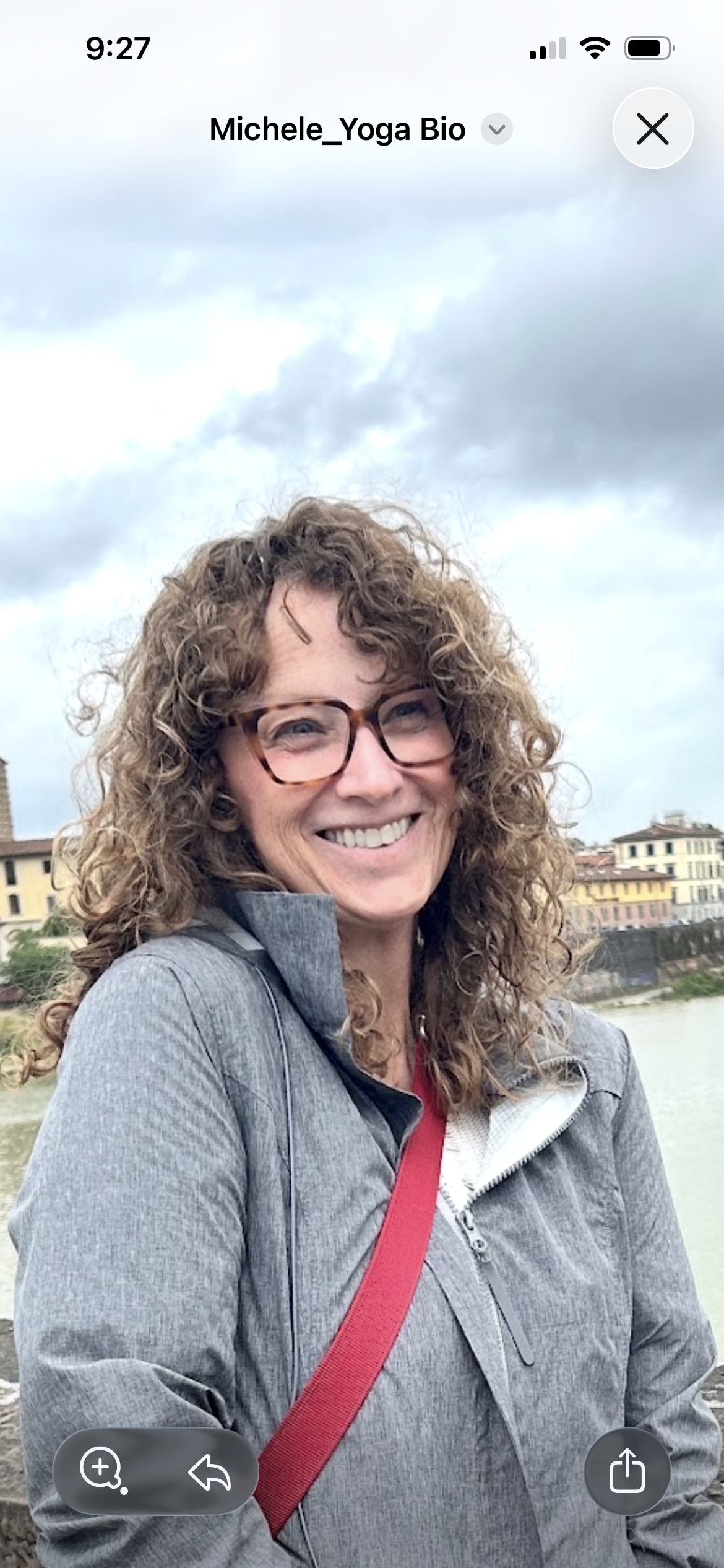 A woman with curly hair and glasses smiling outdoors near a body of water with buildings in the background on a cloudy day.