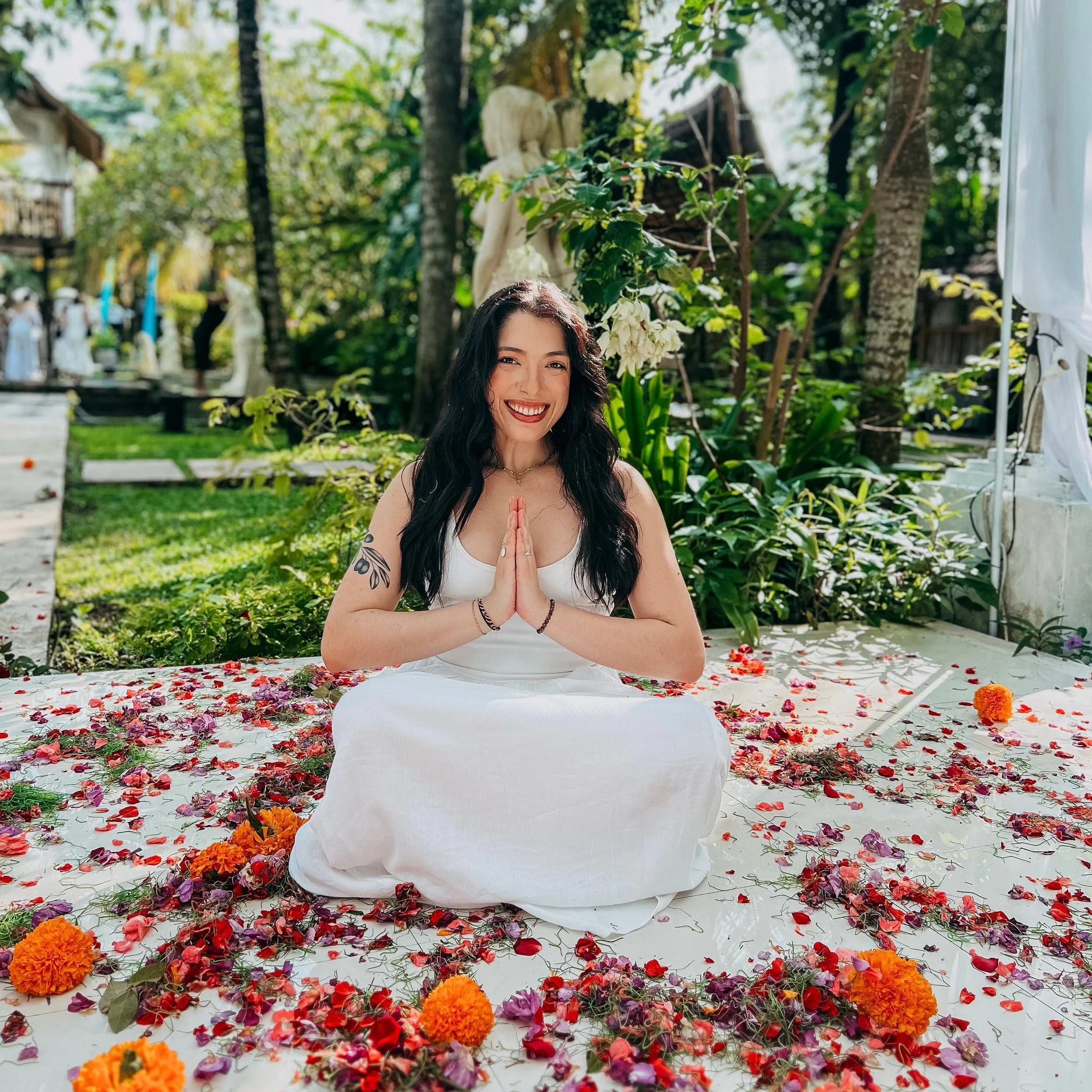 A woman practicing meditation outdoors amid colorful flower petals and marigold flowers, with greenery and trees in the background.