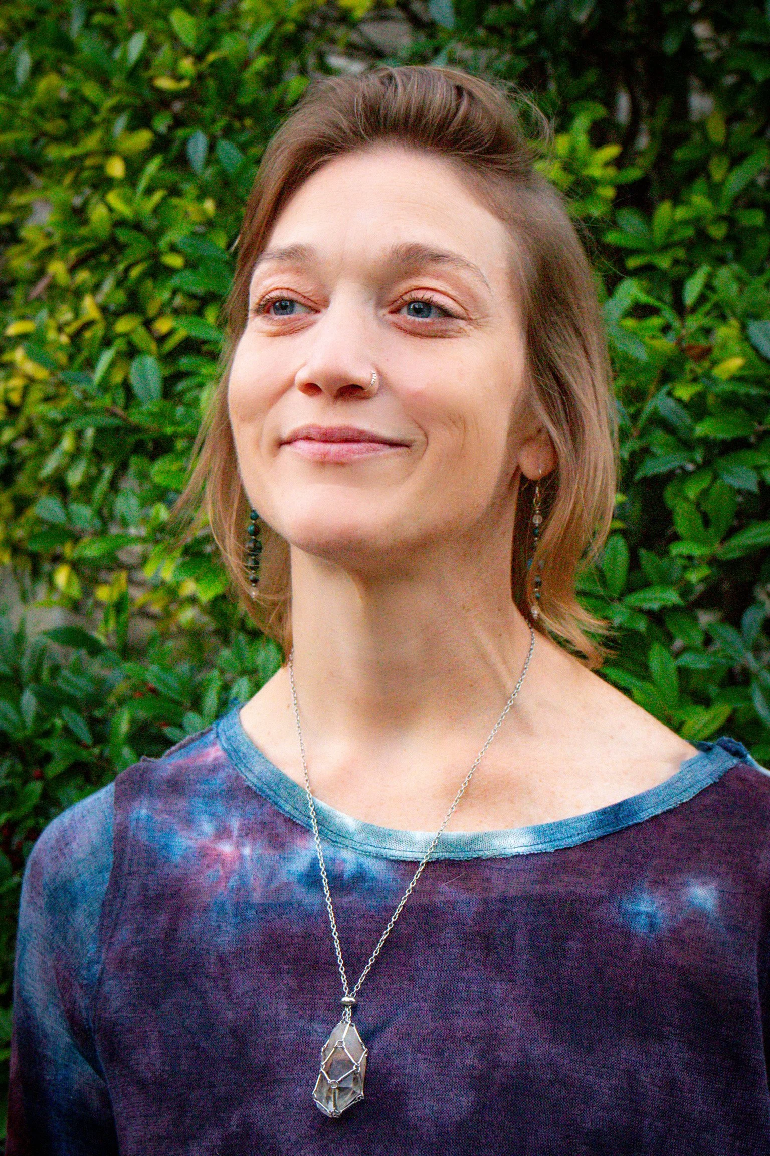 A young woman with short, light brown hair and bright blue eyes stands outdoors in front of green foliage. She is wearing a colorful tie-dye shirt and silver jewelry, including earrings, a necklace, and a large geometric pendant.