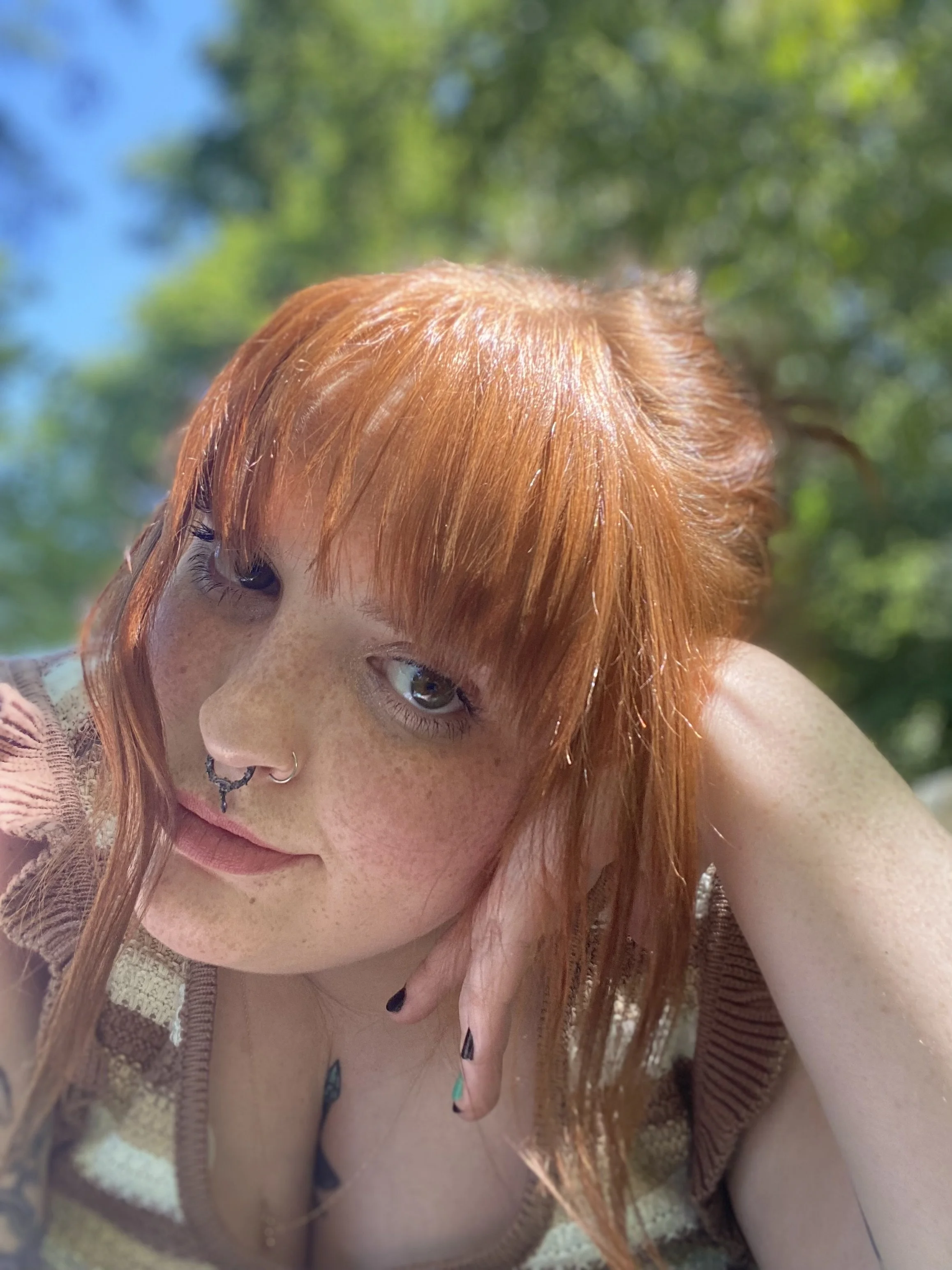 Close-up of a woman with red hair, freckles, and multiple facial piercings outdoors with green foliage background.