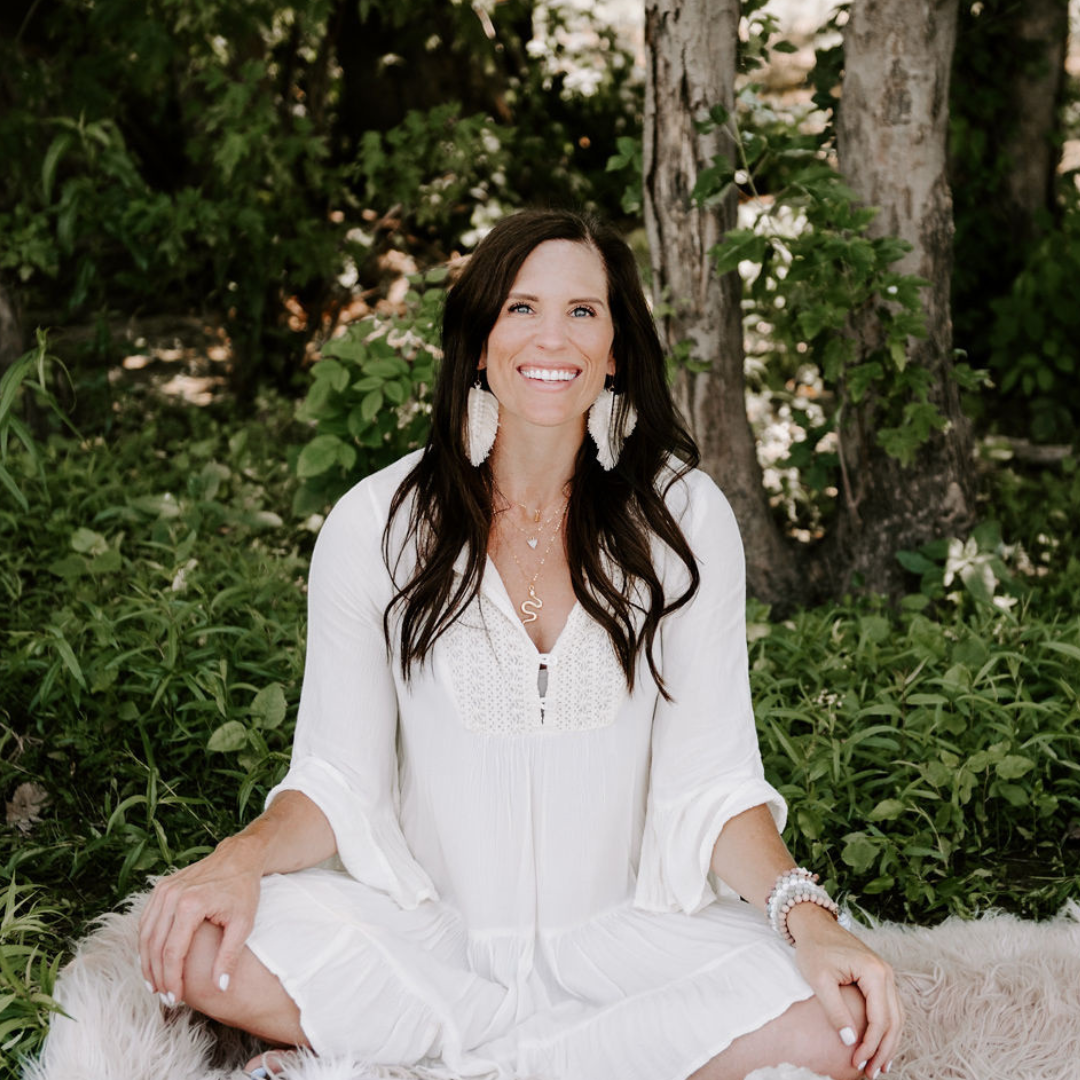 A woman with long dark hair smiling, sitting cross-legged outdoors in front of trees and greenery, wearing a white dress and boho jewelry.