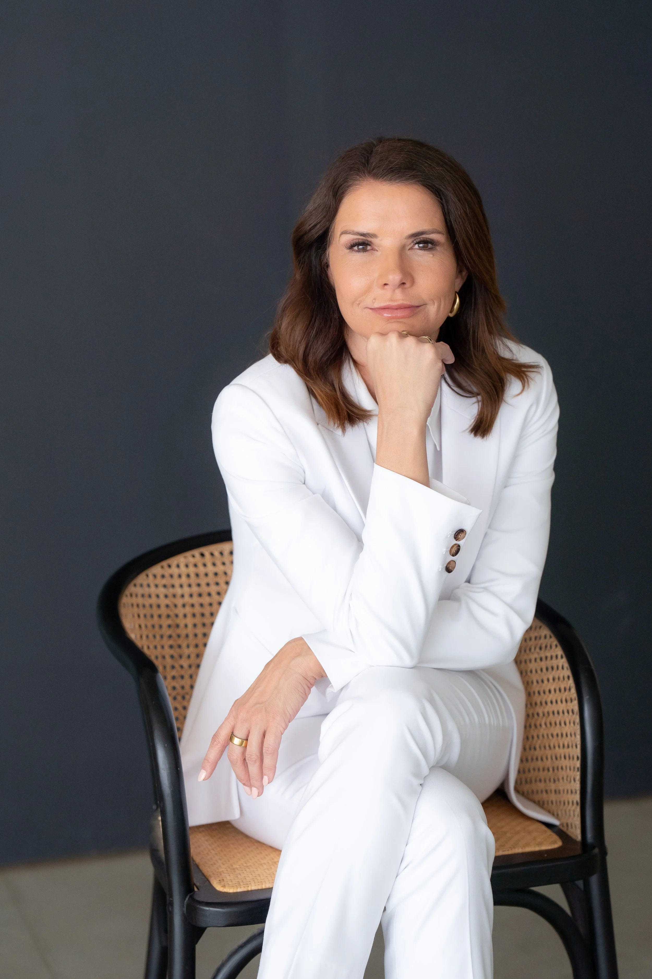 A woman with shoulder-length brown hair and light skin, wearing a white suit, sitting on a wicker chair against a dark gray background, resting her chin on her hand and looking at the camera with a slight smile.