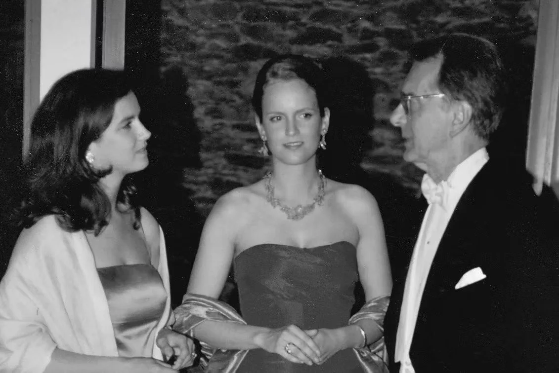 Drei Personen bei einer formellen Veranstaltung, zwei Frauen und ein Mann, alle in eleganter Kleidung, vor einem Hintergrund aus Steinen oder Mauerwerk.