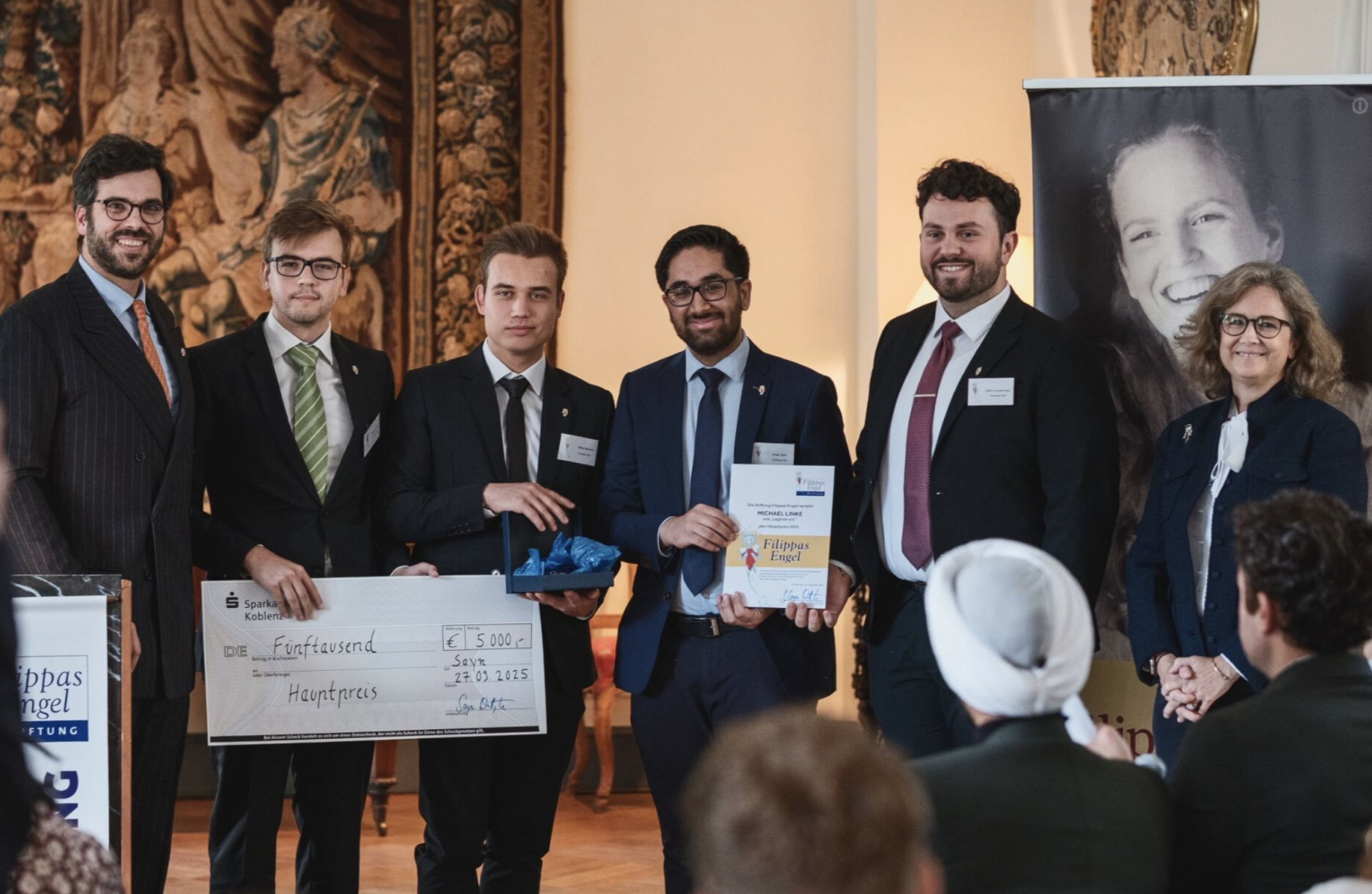 Eine Gruppe von sechs Menschen steht und lächelt bei einer Preisverleihung, während sie eine große Scheck- und eine Urkunde halten.