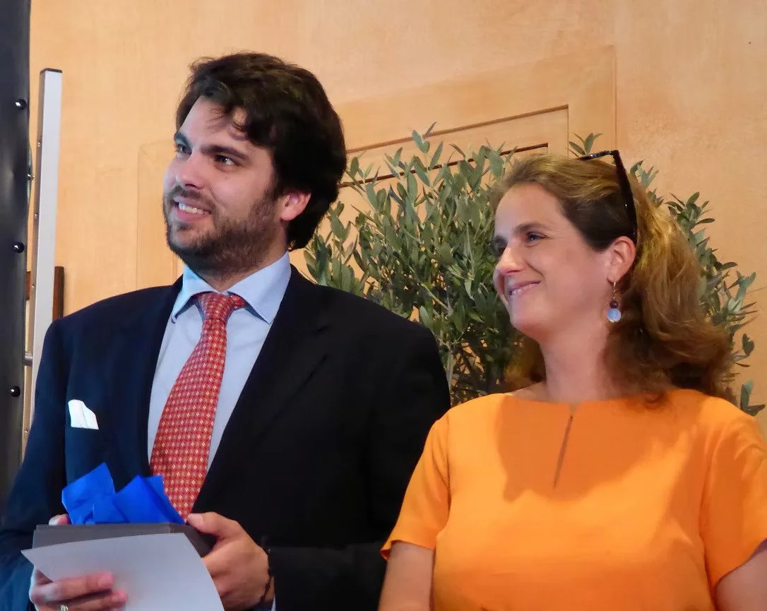 Ein Mann in Anzug und Krawatte steht neben einer lachenden Frau in Orange, im Hintergrund eine Pflanze.