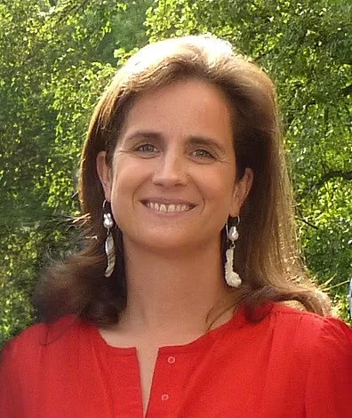 Eine lächelnde Frau mit braunen Haaren, trägt eine rote Bluse und große Ohrringe, im Grünen bei Tageslicht.