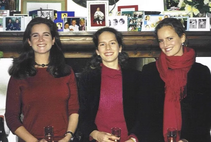 Drei junge Frauen stehen nebeneinander vor einer Holzverkleidung, lachen und halten Gläser mit Getränken. Im Hintergrund eine Wand mit Fotos und Grußkarten.