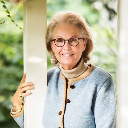 Ältere Dame mit Brille, grauem Pullover und Bluse, steht draußen vor einem Haus mit grüner Natur im Hintergrund.