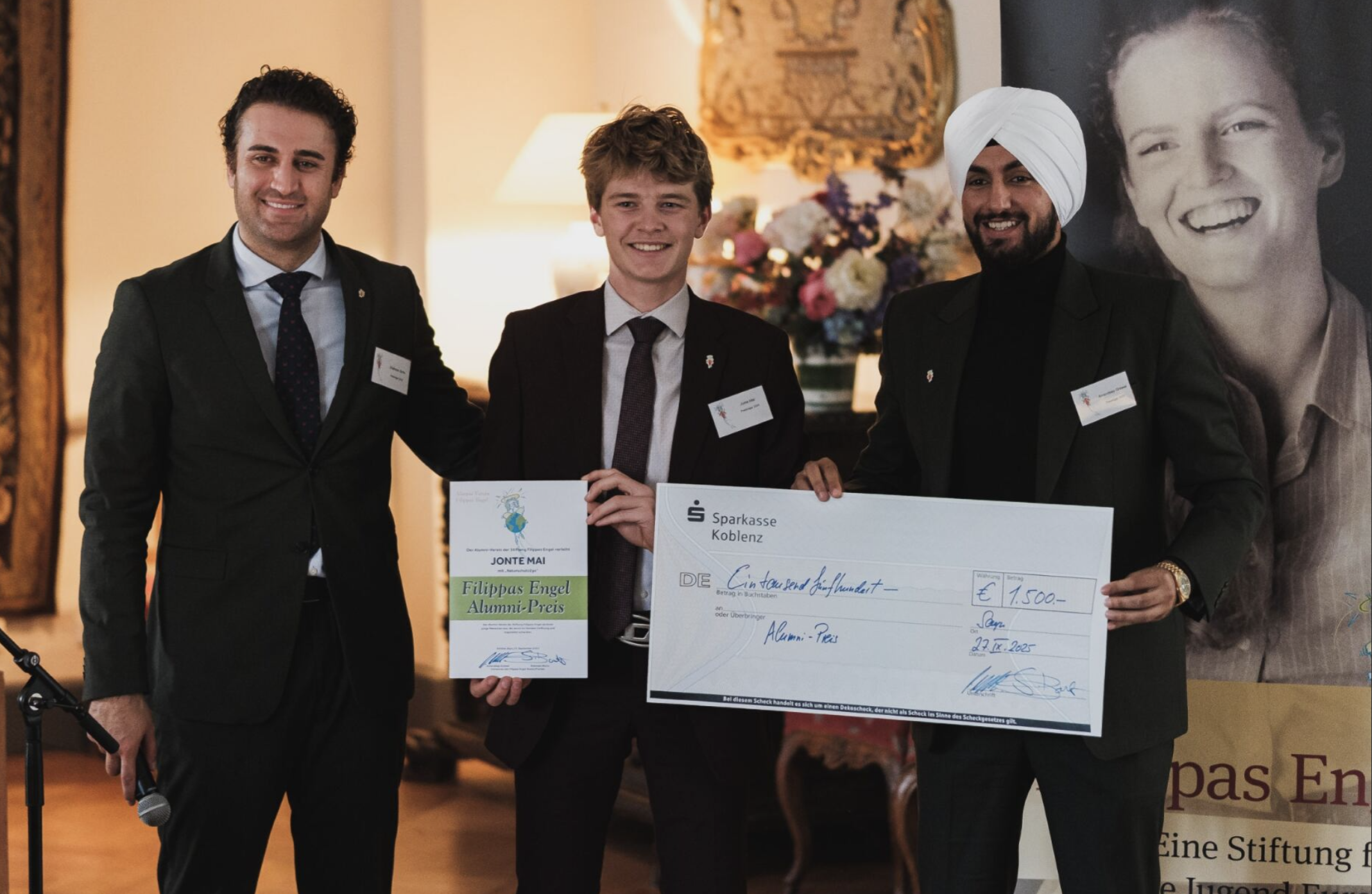 Drei Männer in formeller Kleidung stehen zusammen, halten einen großen Scheck und ein Zertifikat bei einer Preisverleihung. Im Hintergrund sind Blumen und ein großporträt einer lächelnden Frau zu sehen.