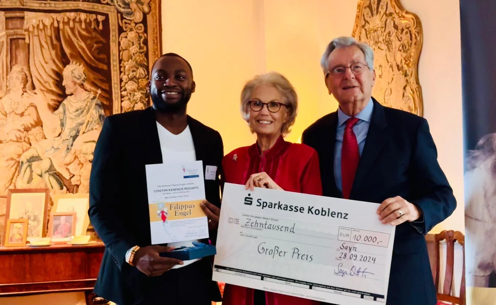 Drei Menschen, die bei einer Preisverleihung eine große Scheck- und Urkunde präsentieren. Hintergrund mit Wandteppich und Bildern.