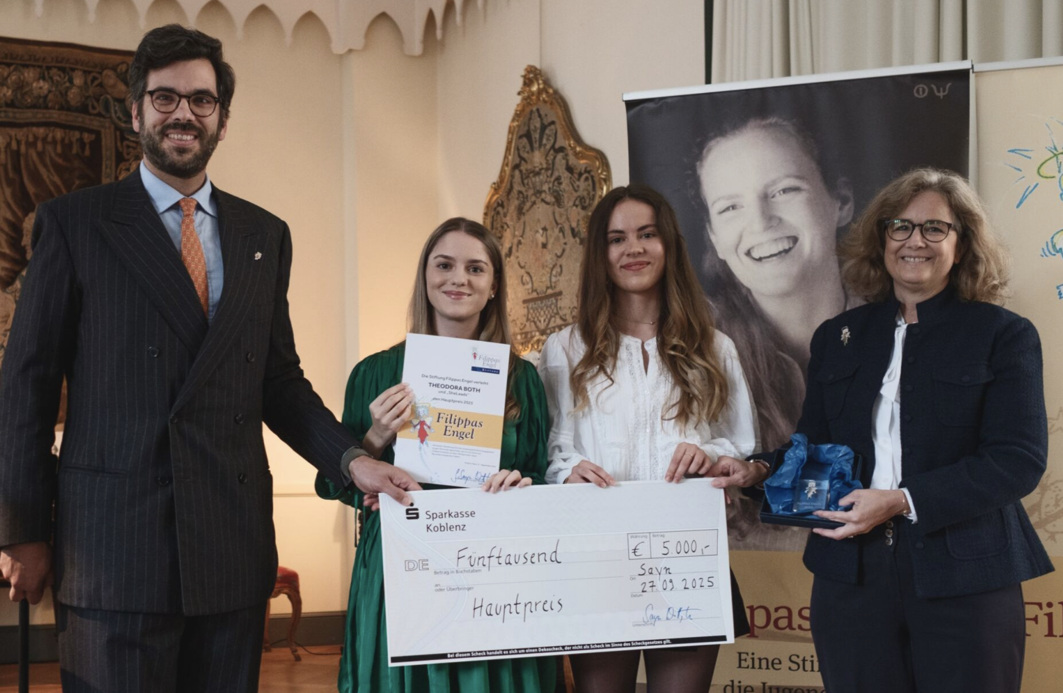 Eine Gruppe von fünf Personen bei einer Preisverleihung, mit einem großen Scheck in der Hand. Im Hintergrund ein schwarz-weiß Porträt einer lachenden Frau, eine Trophäe und ein Gemälde.