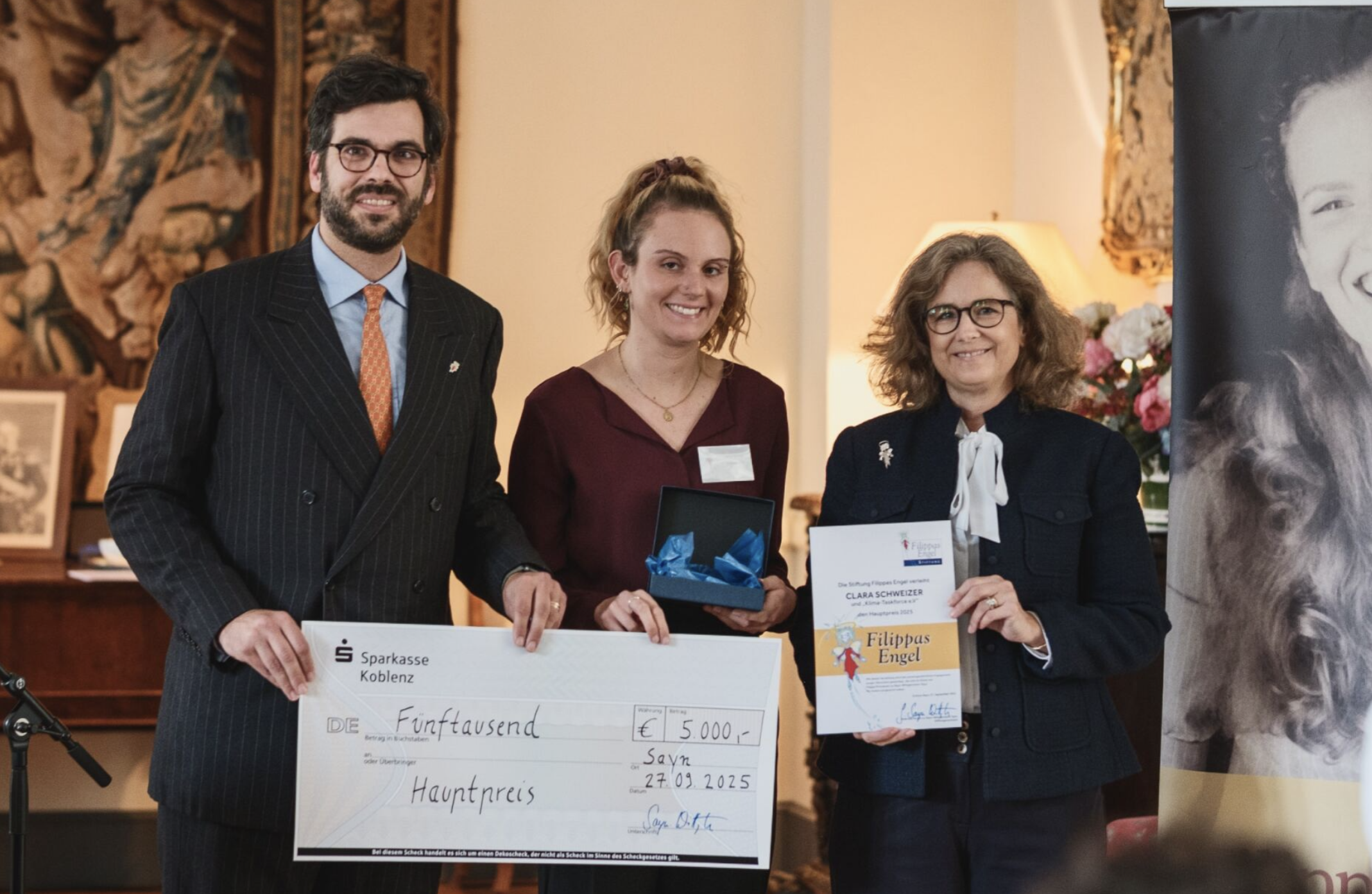Drei Personen bei einer Preisverleihung. Eine Person hält einen großen Scheck im Wert von 5000 Euro, eine andere hält eine Urkunde, alle drei stehen vor einem Gemälde.