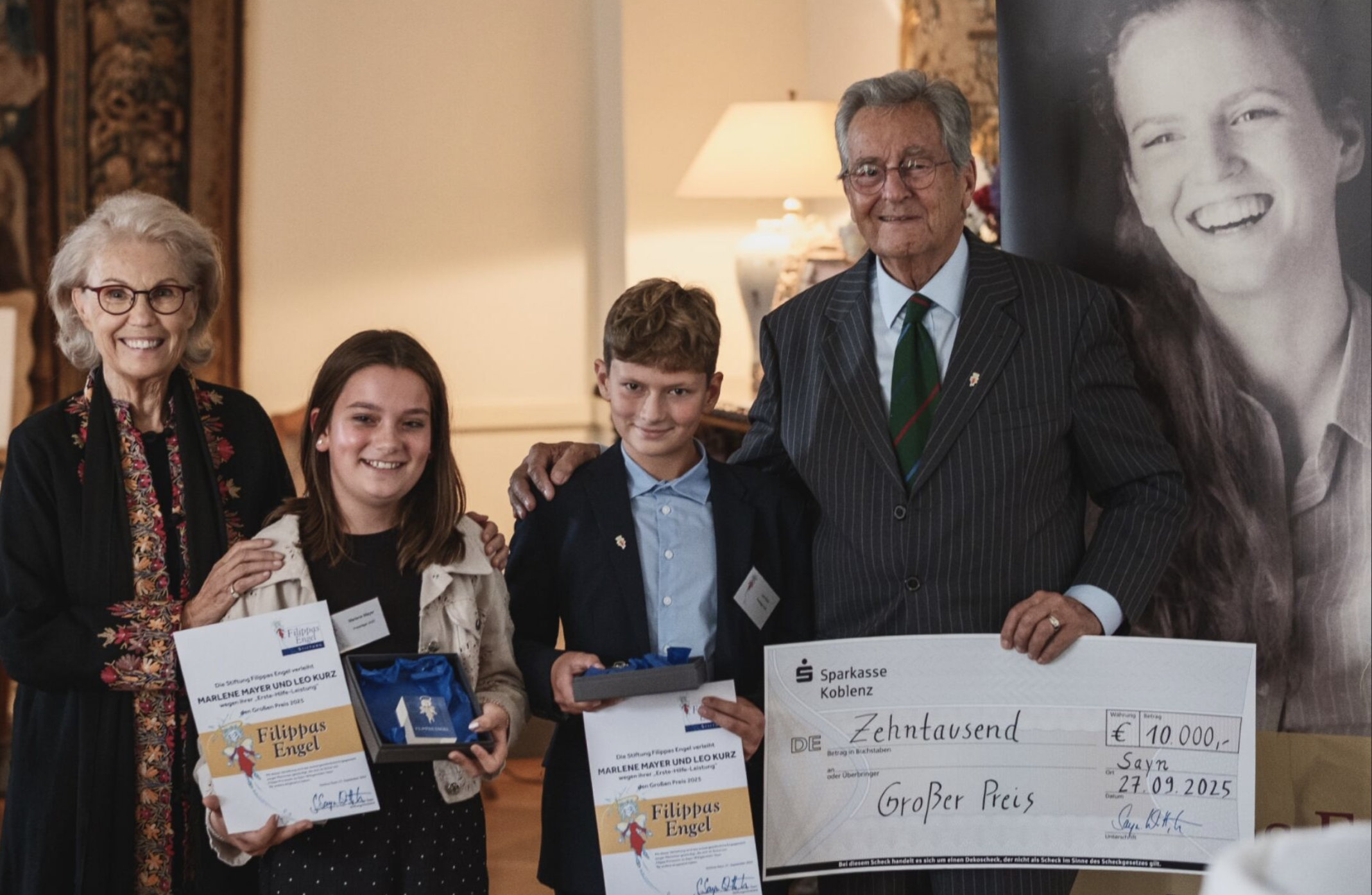 Eine Gruppe von fünf Menschen, bestehend aus älteren und jüngeren Personen, posiert bei einer Preisverleihung. Sie halten Urkunden, Geschenke und einen großen Scheck über 10.000 Euro. Im Hintergrund befinden sich einige dekorative Elemente und ein großer Schwarz-Weiß-Fotodruck einer lachenden Frau.