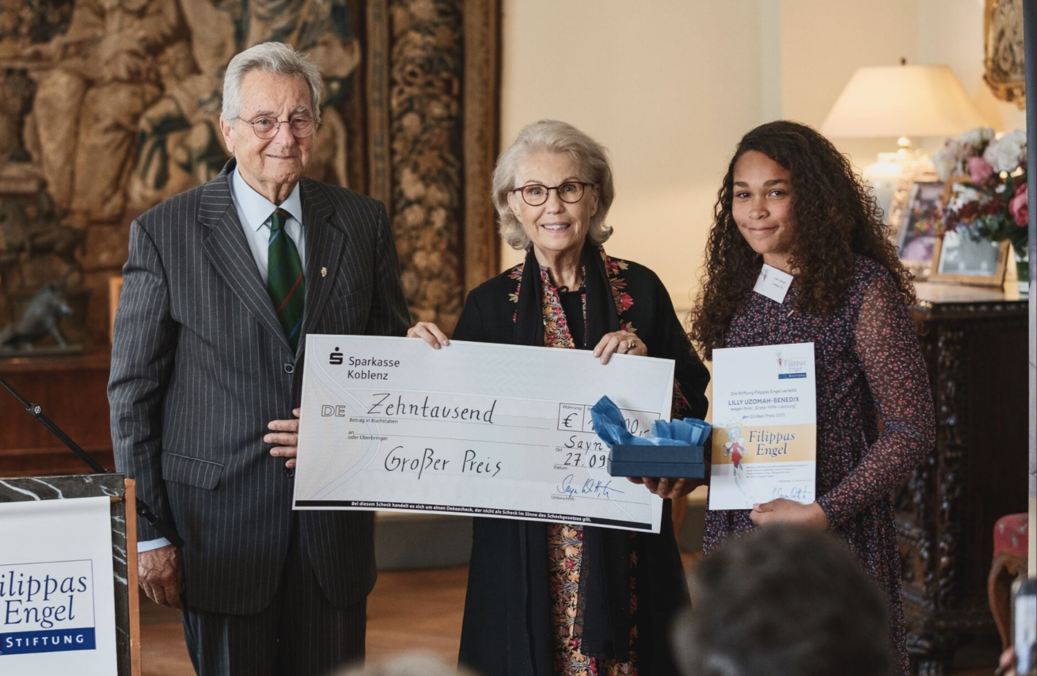 Drei Personen bei einer Preisverleihung halten einen großen Scheck mit der Aufschrift ‚Zehntausend’ sowie eine Tafel, während sie lächeln. Im Hintergrund sind ein Wandteppich, eine Lampe und Blumen zu sehen.