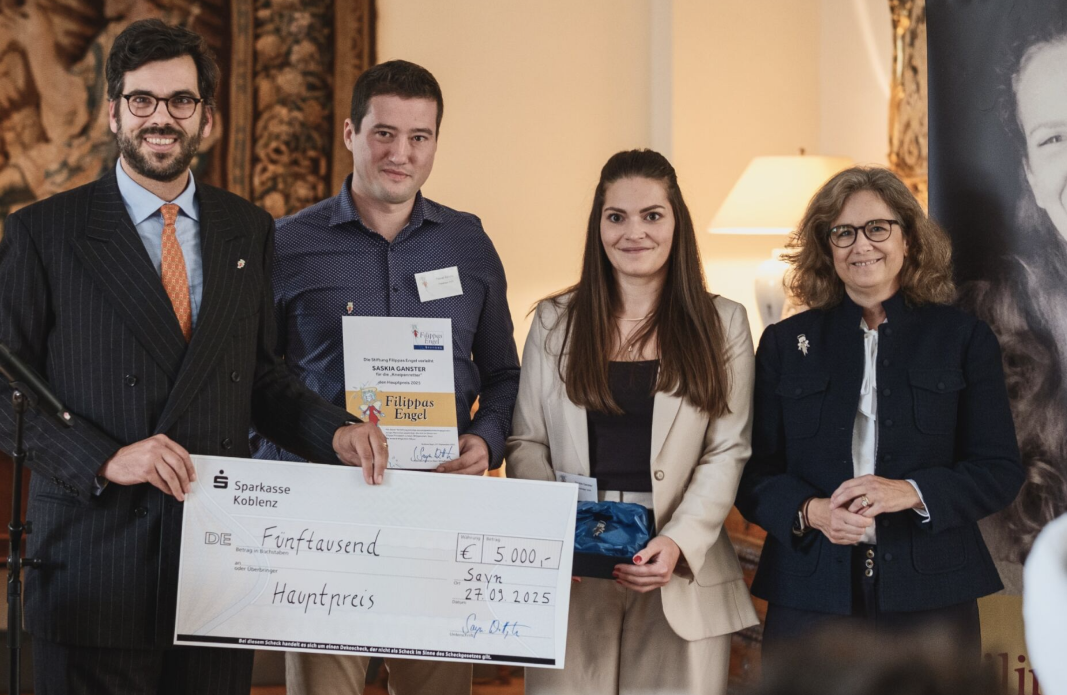 Vier Personen stehen in einem Raum und präsentieren einen großen Scheck, auf dem 5000 Euro stehen. Es handelt sich um eine Preisverleihung oder Spendenübergabe, mit einem festlichen Hintergrund und einem großen Porträt an der Wand.