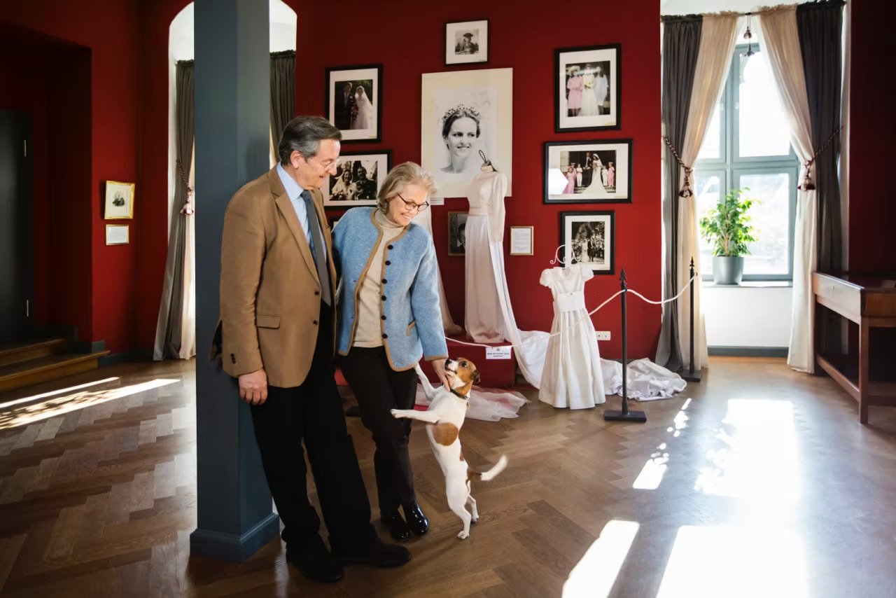 Ein älteres Paar hält Händchen und spielt mit einem Hund in einem Museum, während sie Fotos und Kleiderversionen auf einer roten Wand betrachten.