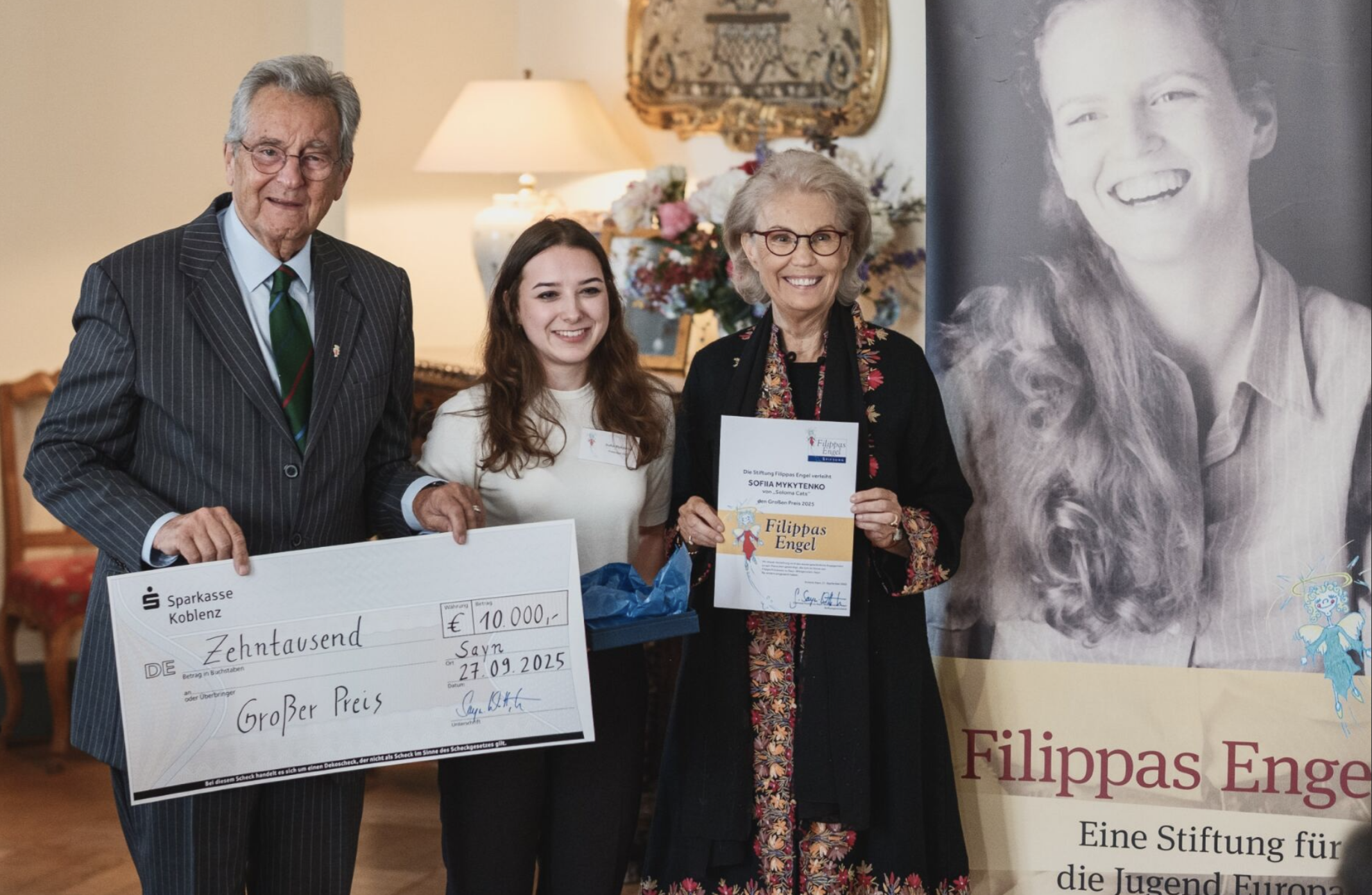Eine Gruppe von vier Menschen beim Preisverleihung, die eine große Scheck und eine Urkunde halten, im Hintergrund ein Bild eines lachenden Jungen und Blumen.