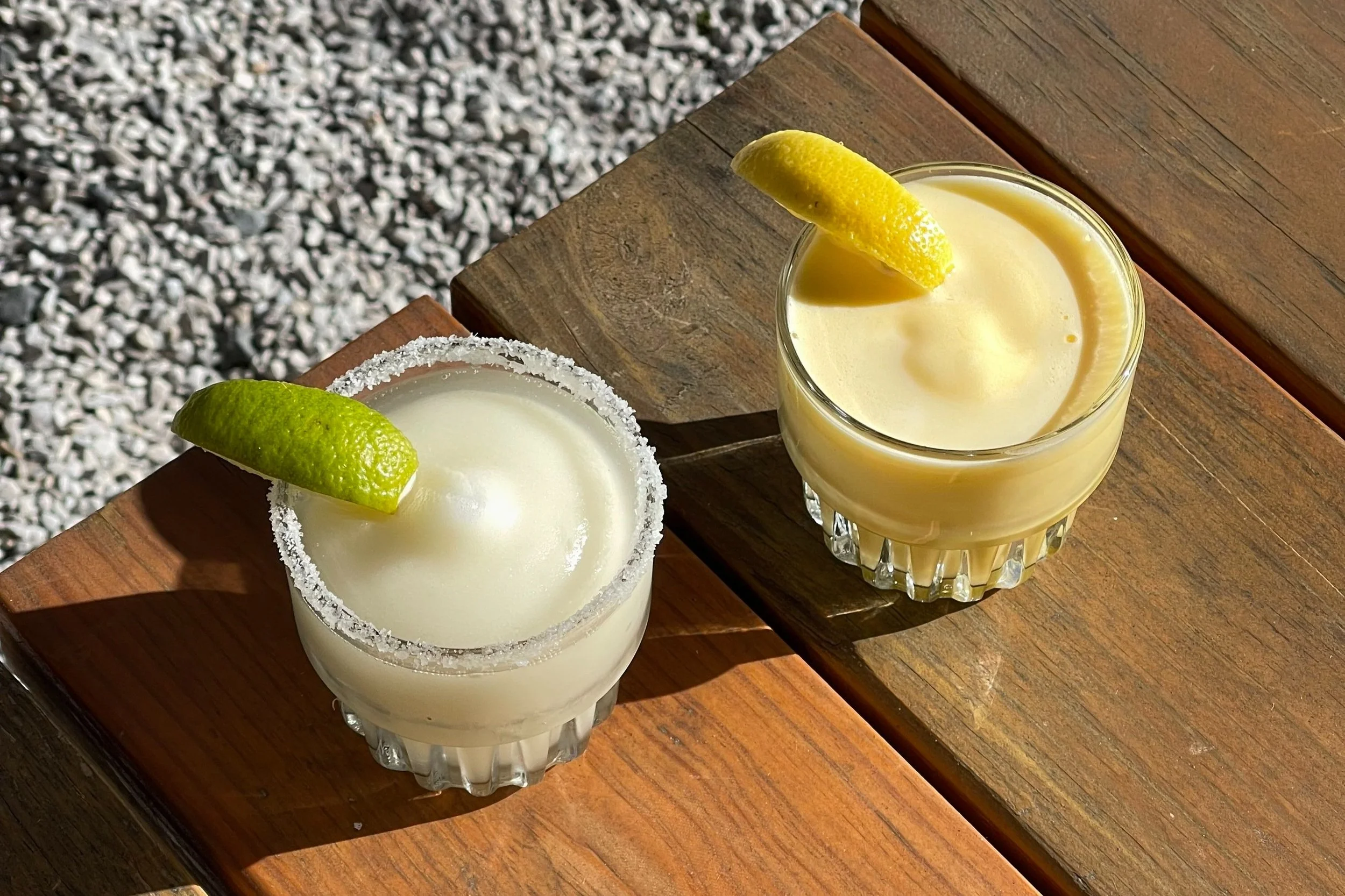 Two glasses of frozen margaritas with lime wedges, one rimmed with salt, on a wooden table outdoors.
