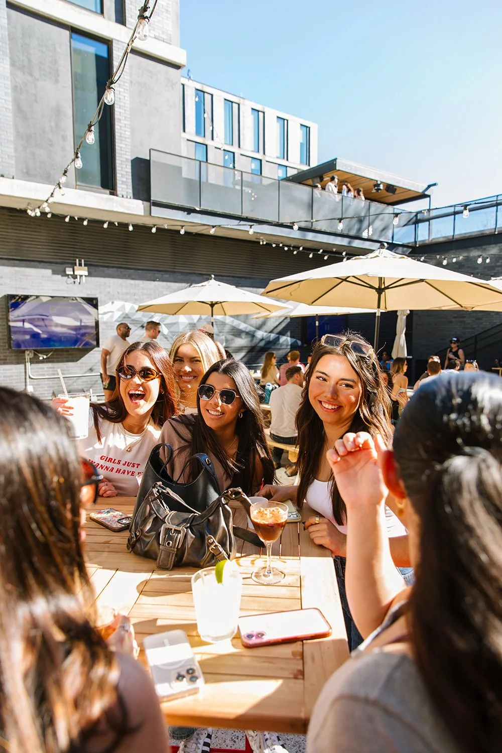 A group of friends at an outdoor patio restaurant enjoying drinks and conversation on a sunny day, with umbrellas and modern buildings in the background.