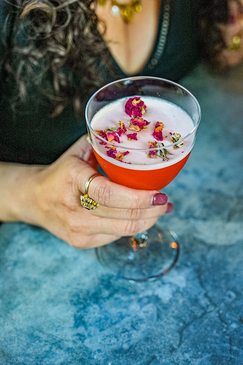 A person with dark curly hair, wearing a black top, holding a stemmed glass filled with a pink cocktail garnished with dried rose petals.