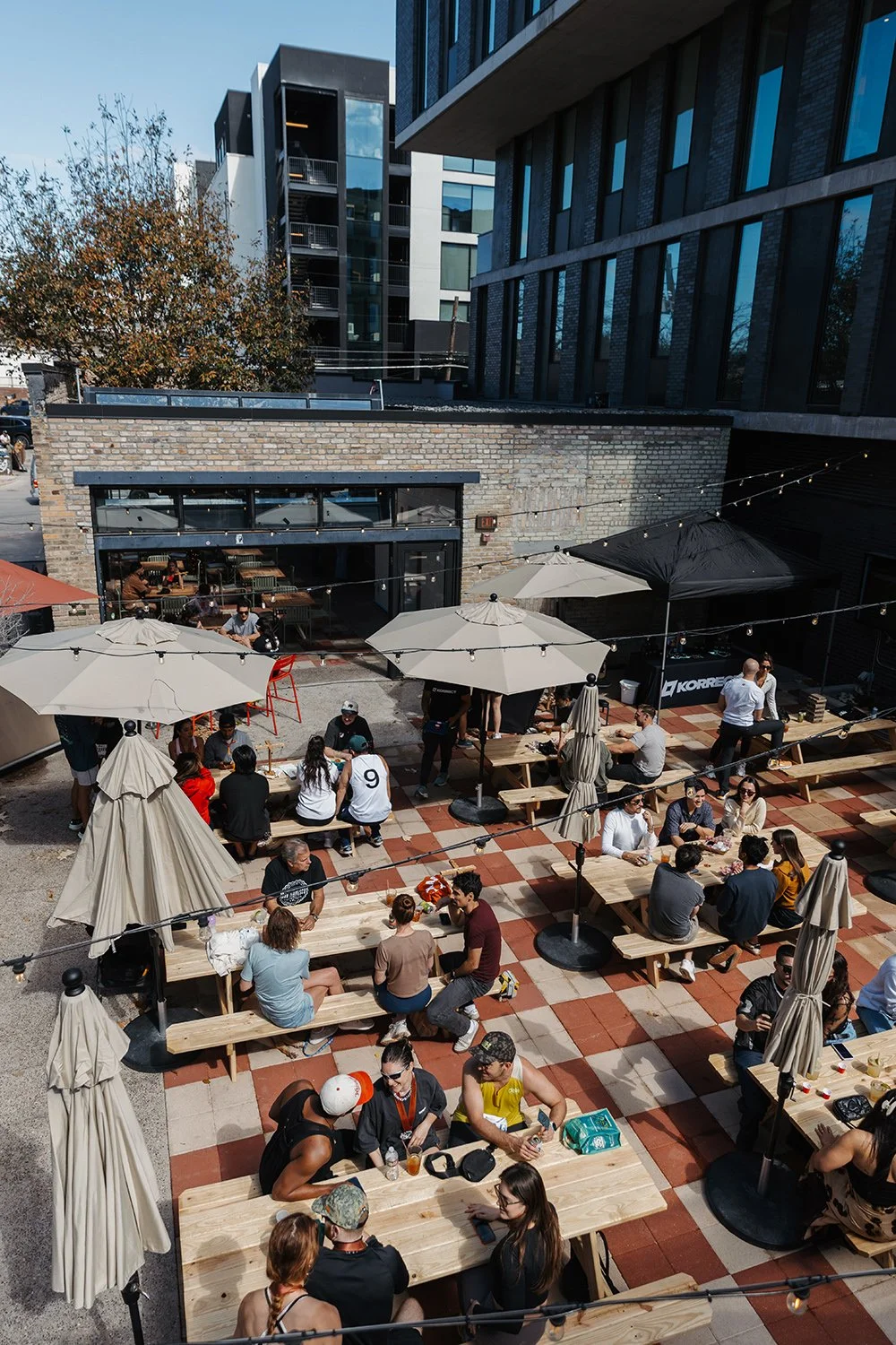 Outdoor patio with wooden picnic tables, umbrellas, and string lights filled with people dining, in an urban area with modern buildings in the background.