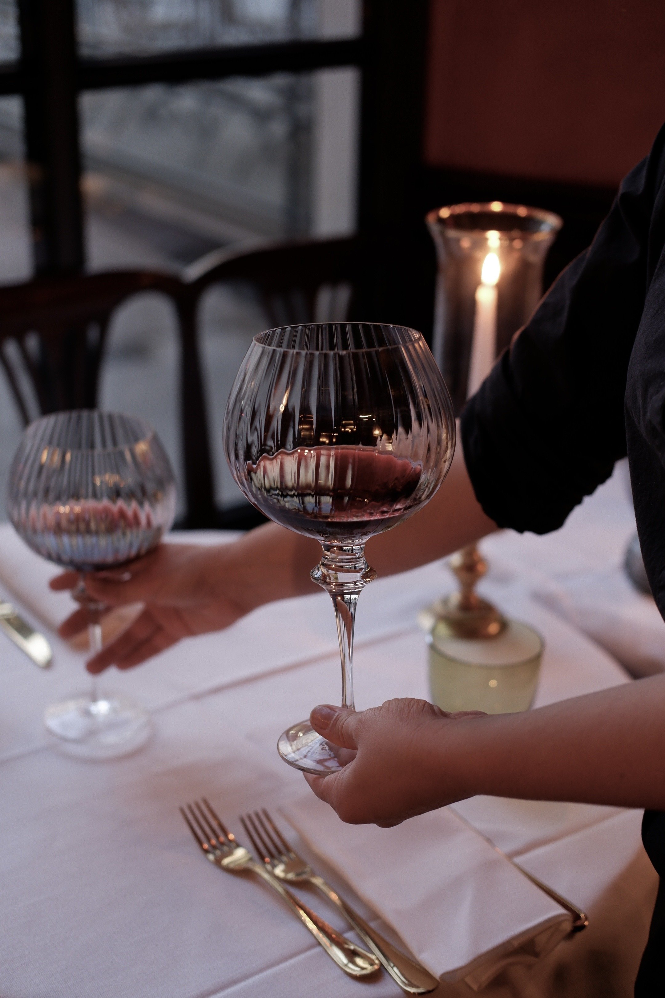 Hands holding wine glasses with red wine at a formal dinner setting, candles on the table in a dimly lit room.