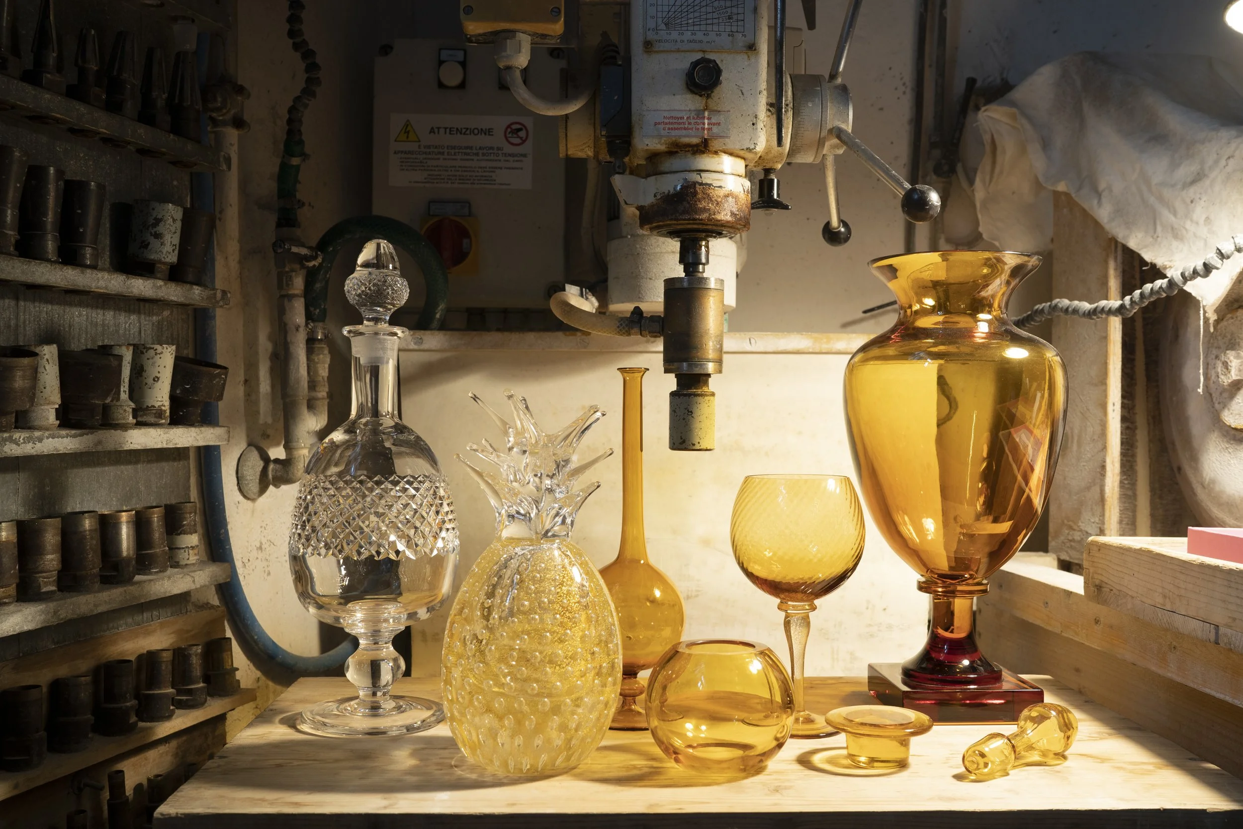 Various glass vases and bowls in shades of amber and clear, arranged on a wooden surface in a workshop or craft room with tools and equipment in the background.