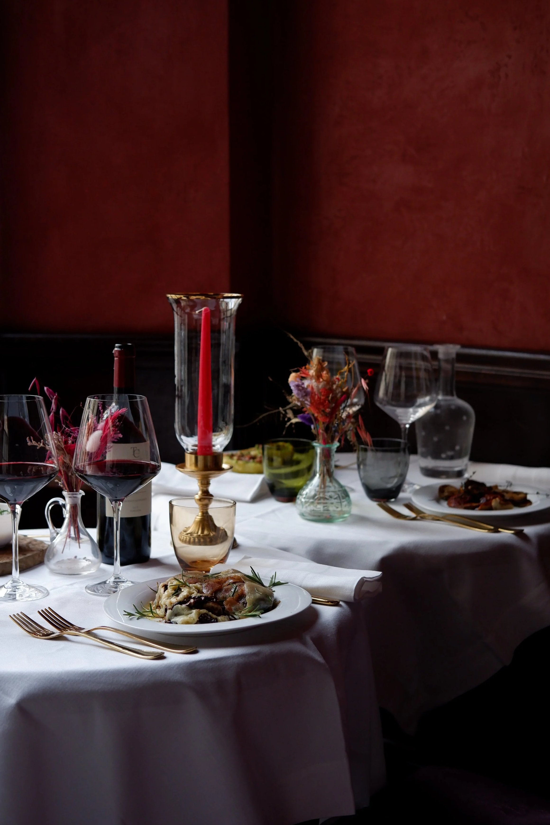 A dining table set for a meal with plates of food, glasses of red wine, and decorative elements like a candle, flowers, and various glassware, with a dark red wall in the background.