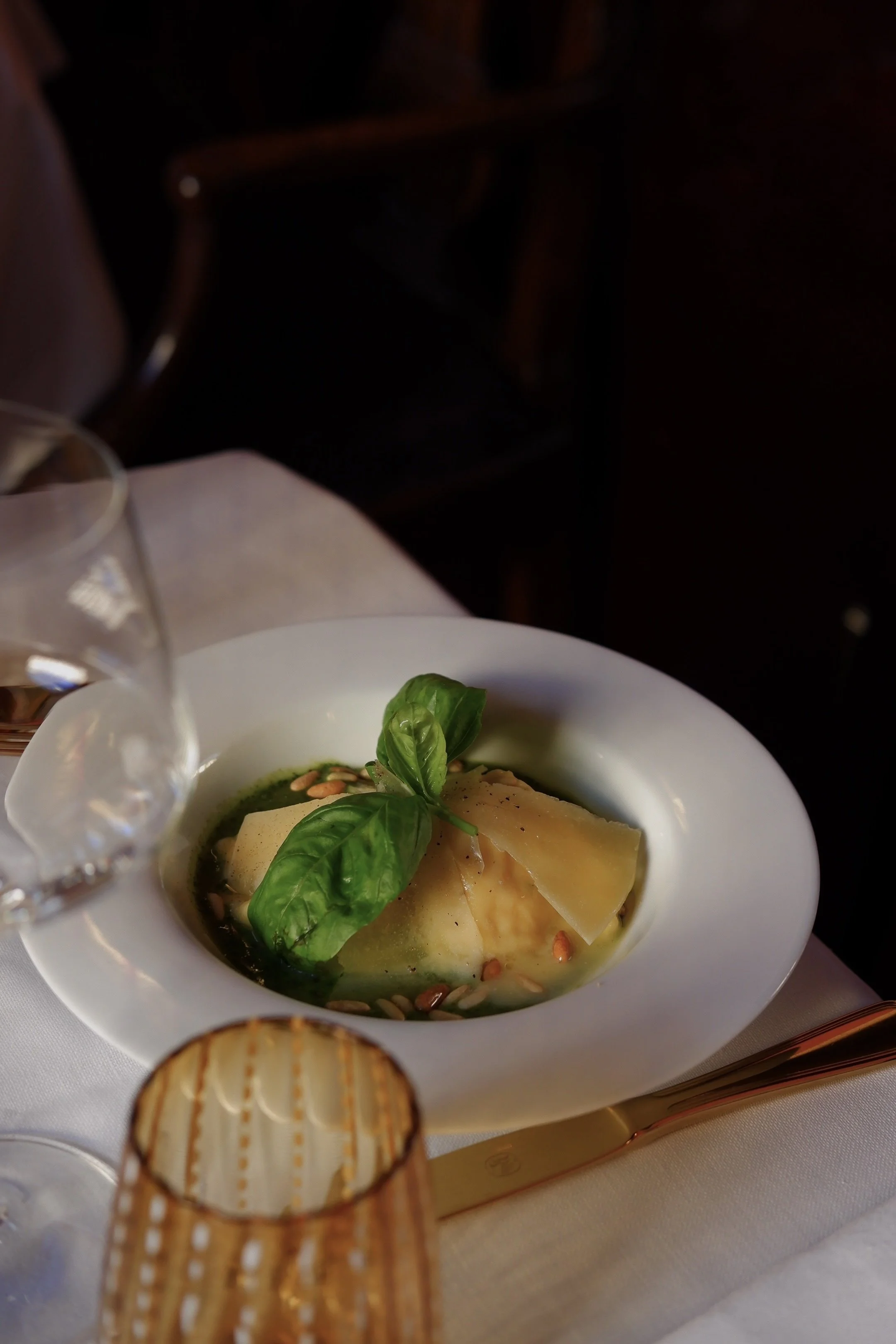 A bowl of Italian pasta topped with fresh basil leaves, cheese, and pine nuts on a white tablecloth, with a glass of water and a gold-colored fork nearby.
