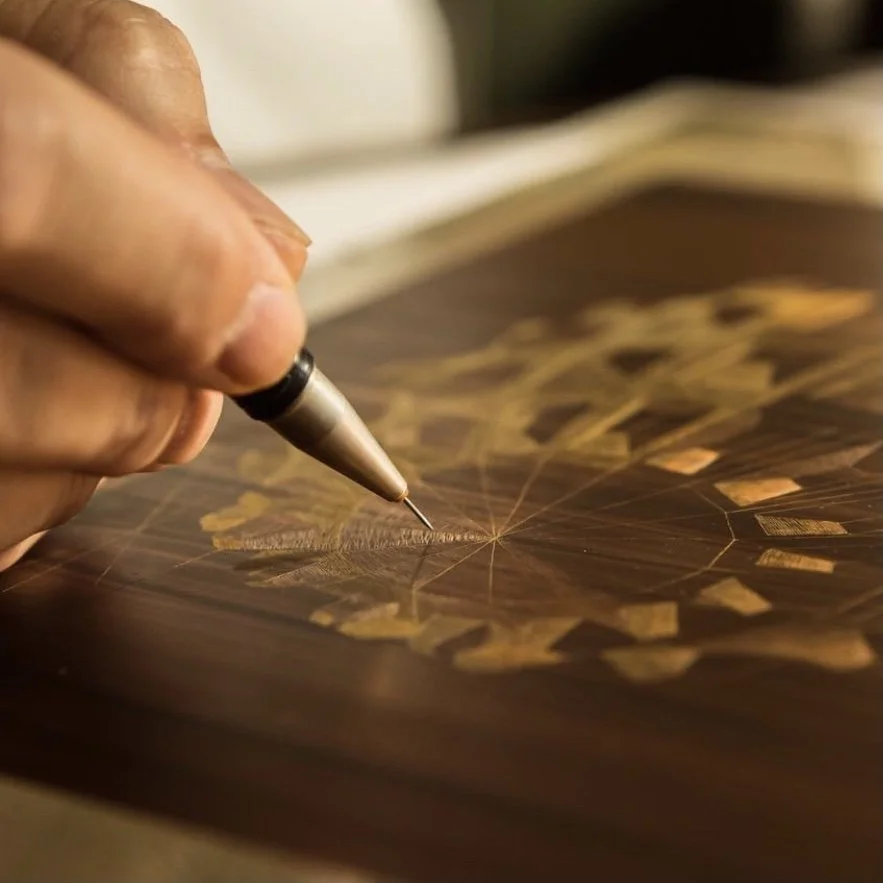 Close-up of a person's hand using a precision tool to carve or etch a detailed geometric design onto a dark wood surface.